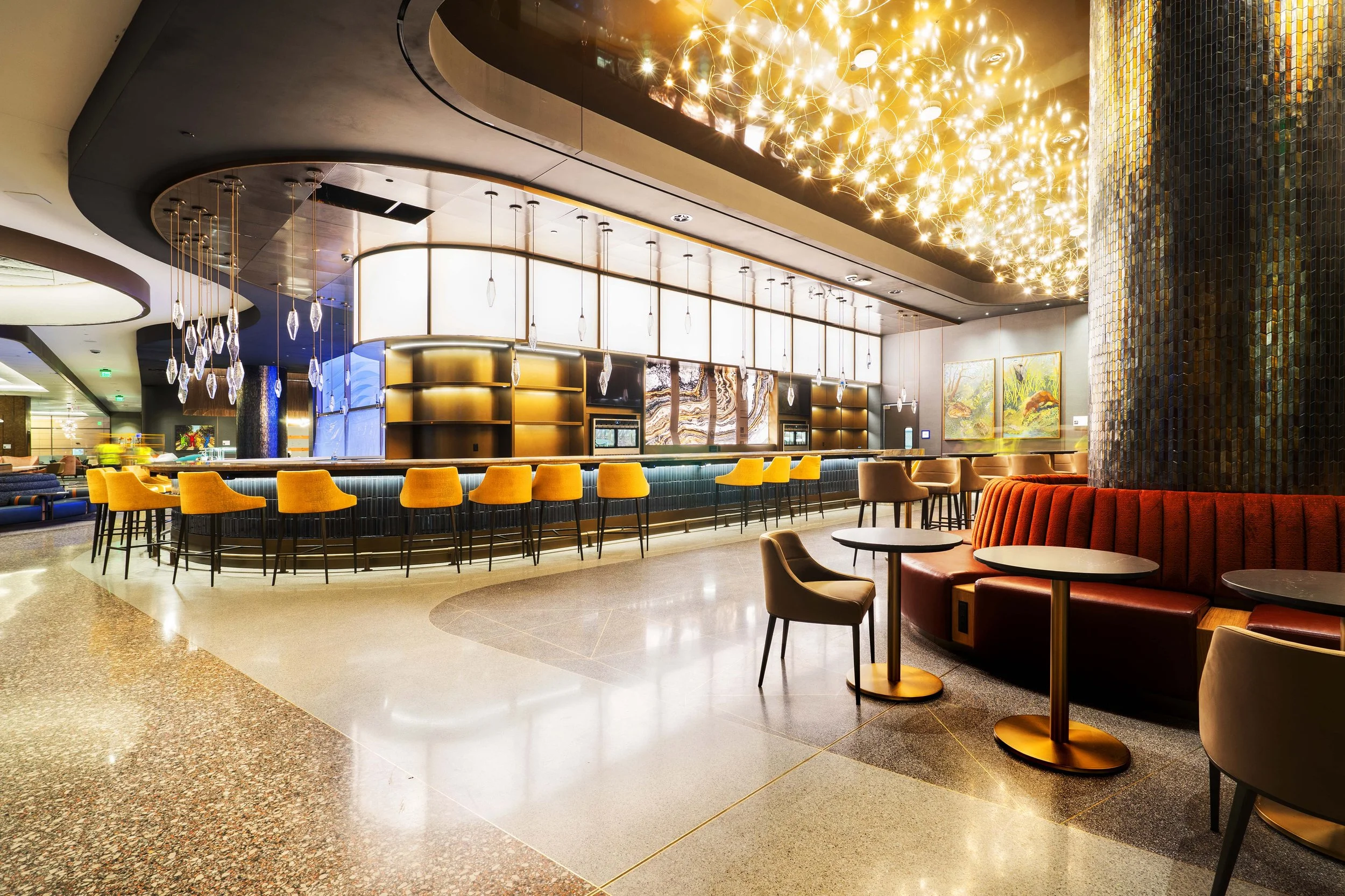 Interior of a modern hotel lobby bar with yellow bar stools, a curved bar counter, ambient lighting, and artwork on the wall.