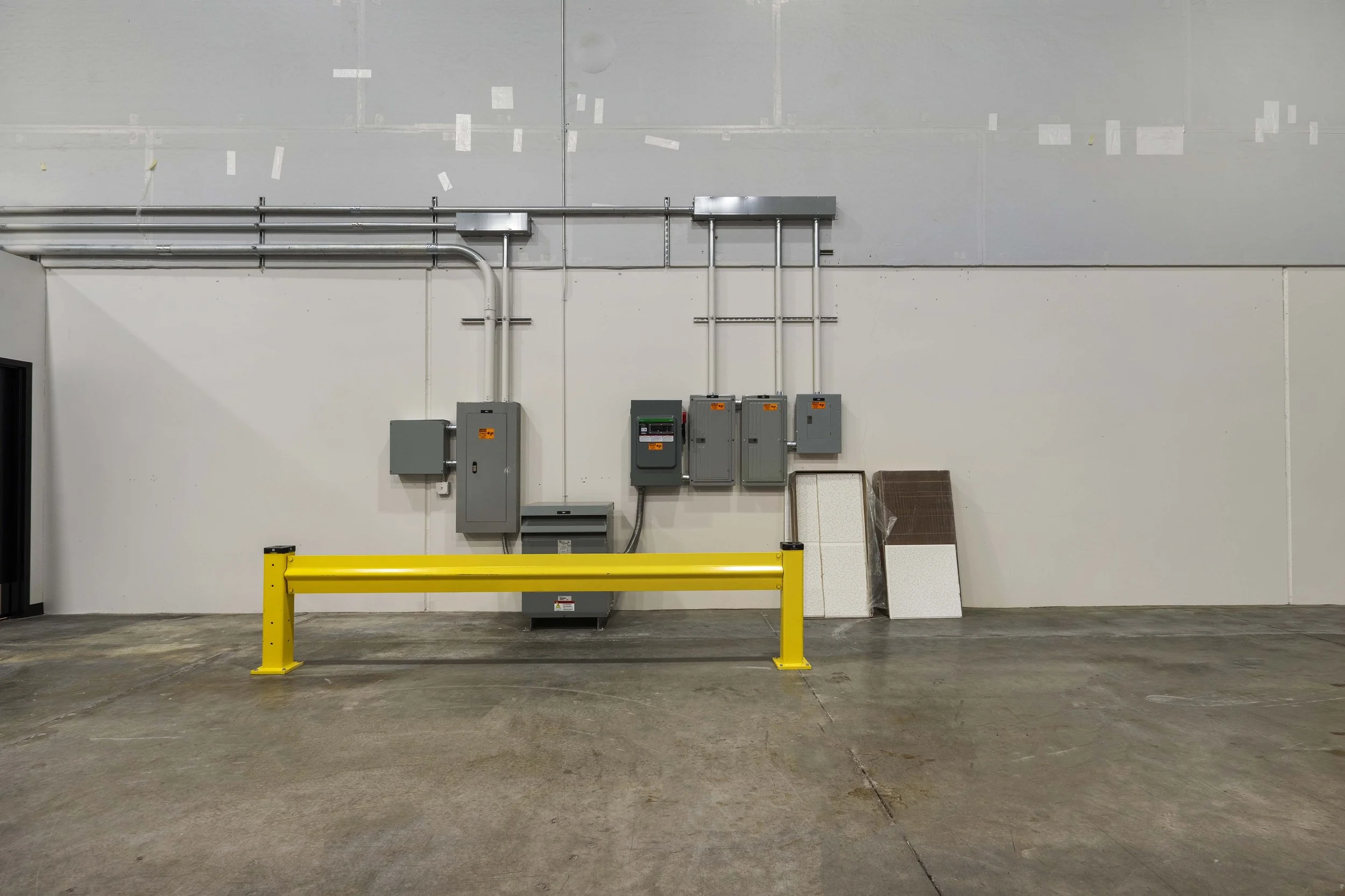 Interior view of an industrial or warehouse space with electrical boxes and gray pipes on the wall, a yellow safety barrier on the concrete floor, and construction panels leaning against the wall.
