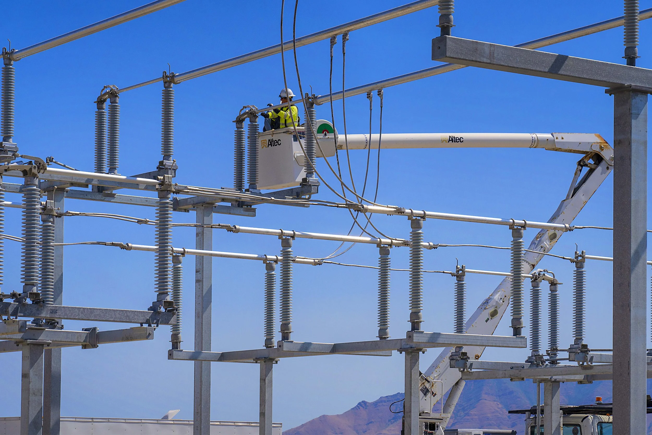 A worker in a safety helmet and vest on a bucket lift repairing an electrical power line against a blue sky, with distant mountains in the background.