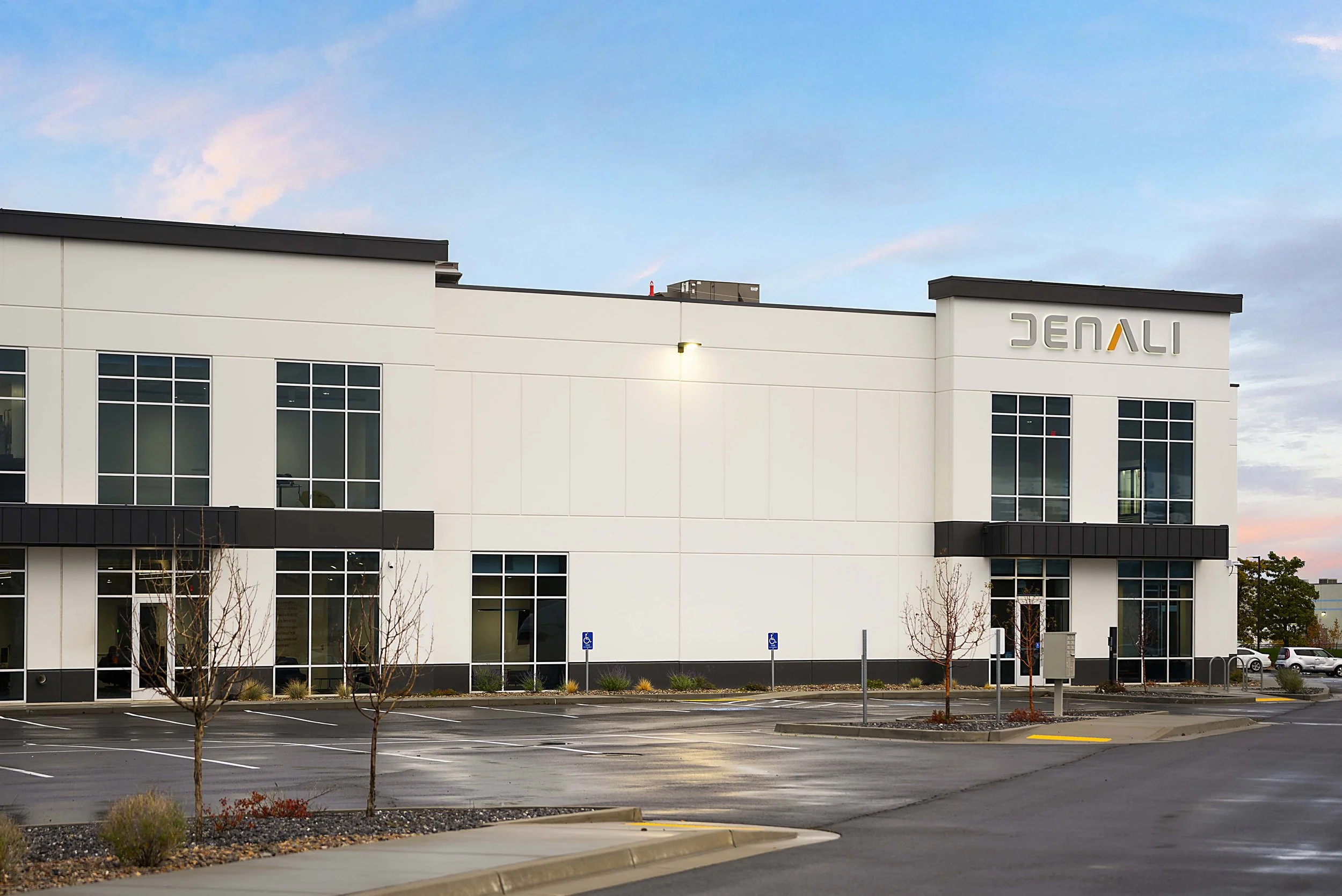 Exterior view of a modern commercial building with the sign 'JENALI' on the top right corner, parked cars, and an empty parking lot with trees and signs for handicapped parking.