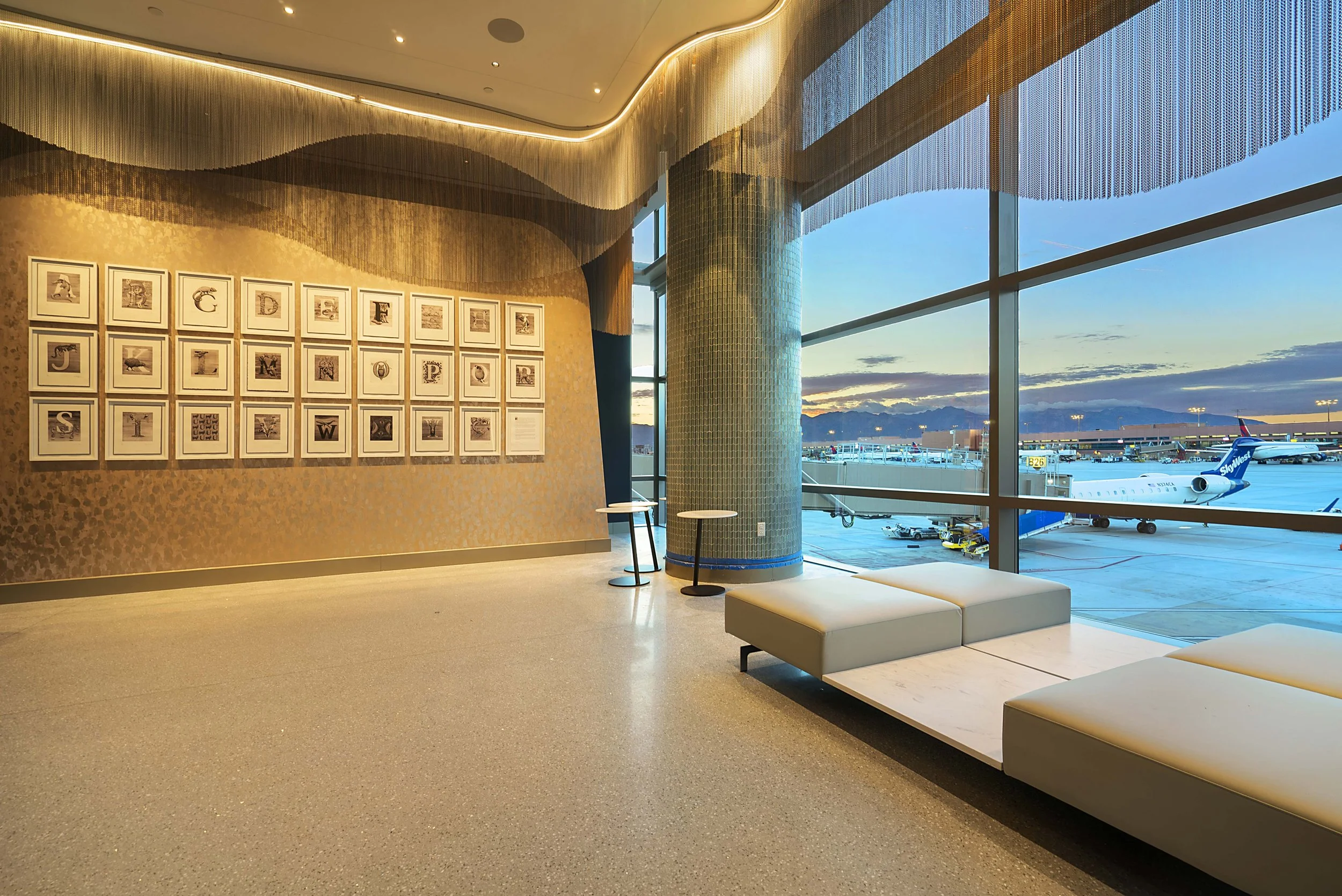 Airport terminal lounge with large viewing windows, modern seating, artwork on the wall, and planes outside at sunset.