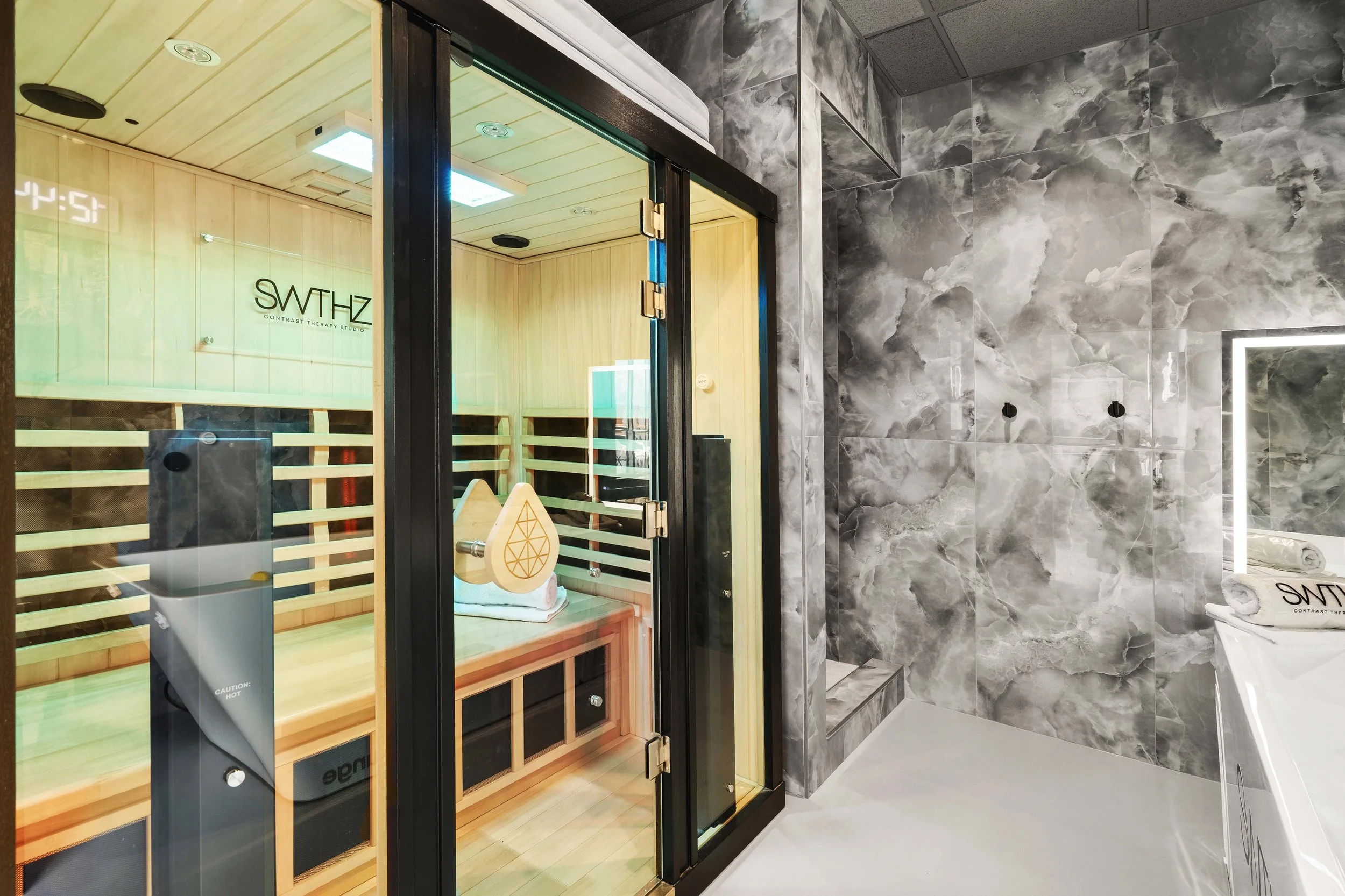 A modern bathroom features a glass-enclosed sauna room with wooden interior and a digital clock on the wall, and a marble tiled shower area with dark gray veined tiles, and a white towel on a heated towel rack.