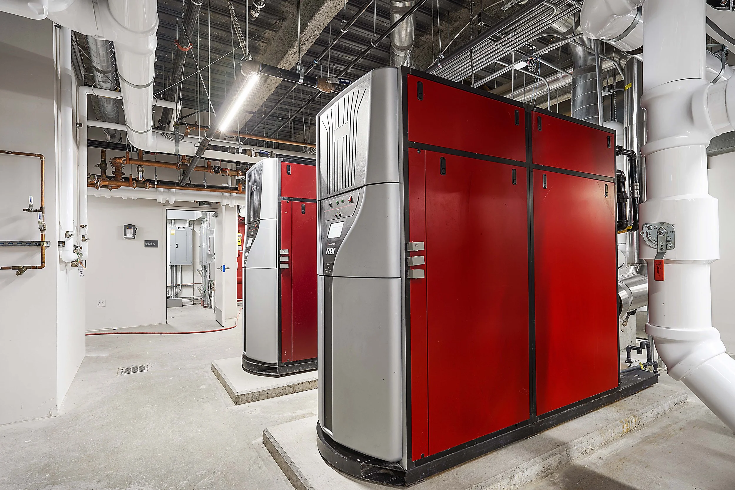 Industrial HVAC or boiler room with large red and gray machinery, white pipes, and exposed ductwork.