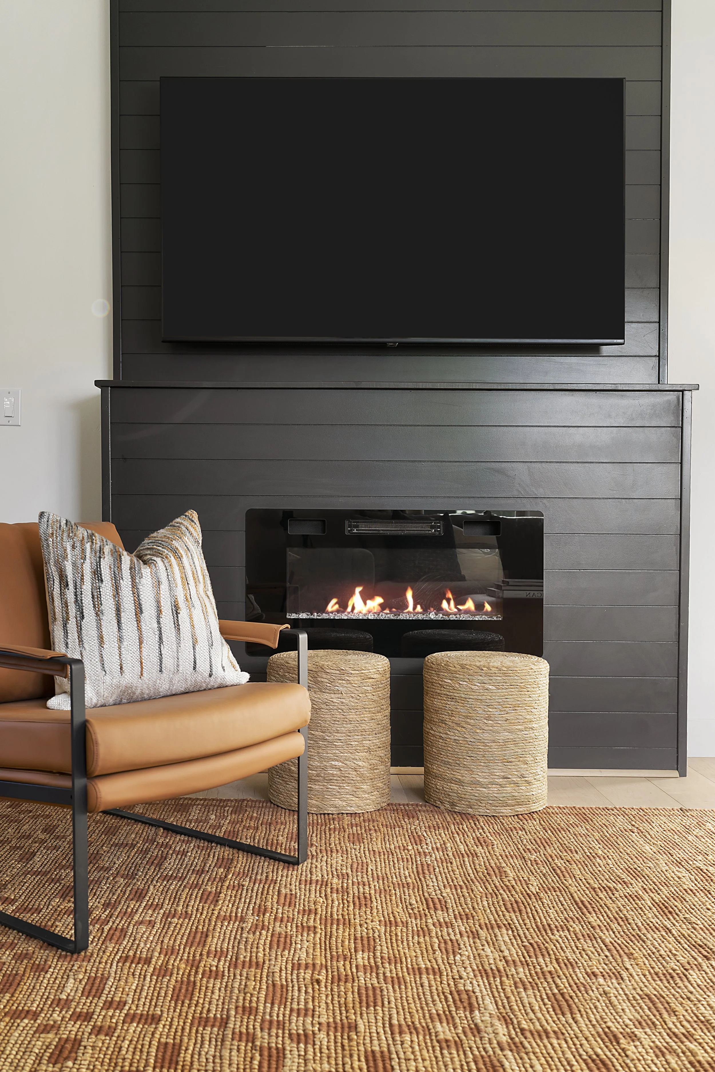 Living room with black-panelled wall, mounted TV, and electric fireplace beneath it. Tan leather armchair with decorative pillow, orange and yellow woven rug, and two round woven stools in front of the fireplace.