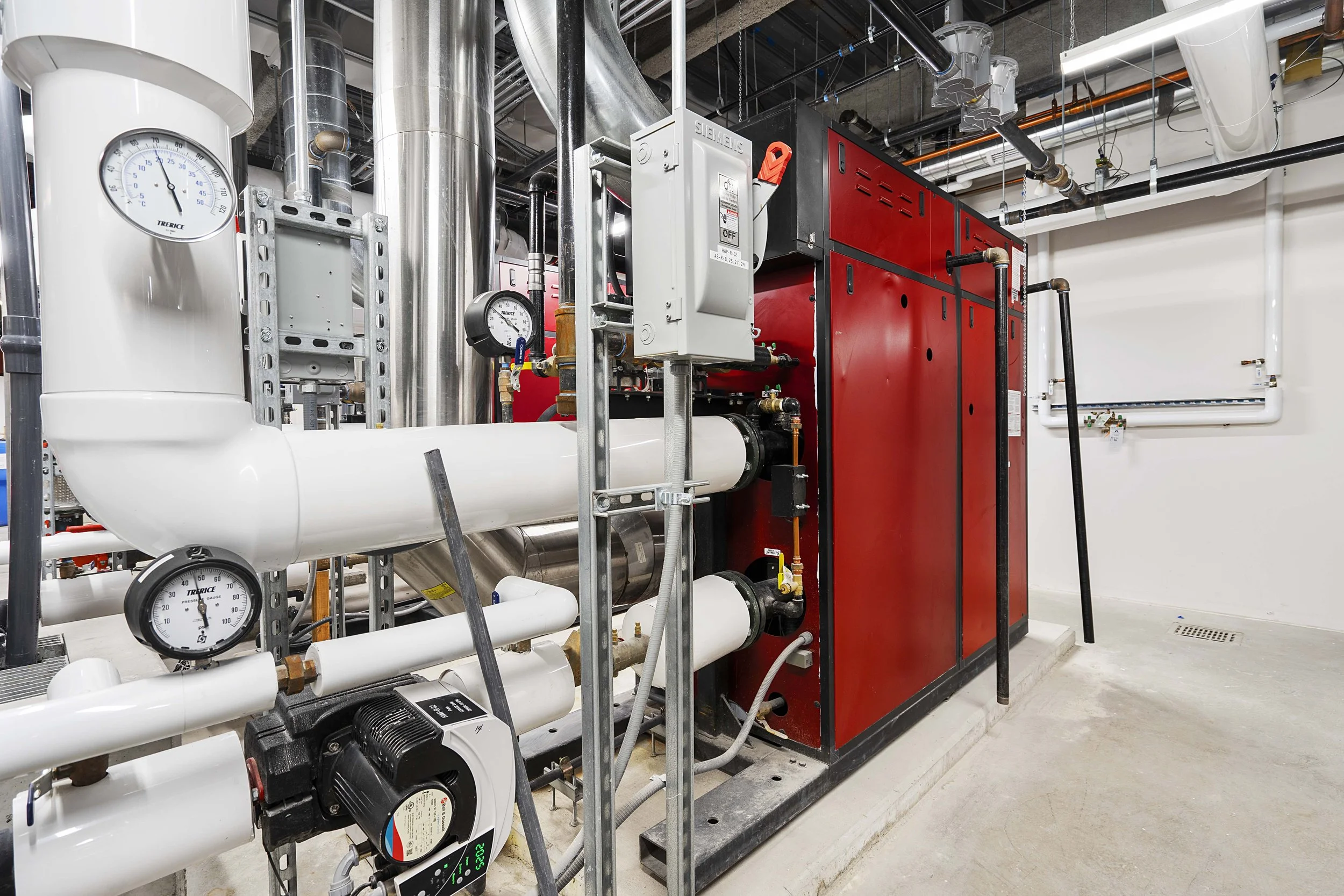 Industrial boiler room with complex pipework, gauges, and a large red heating unit.