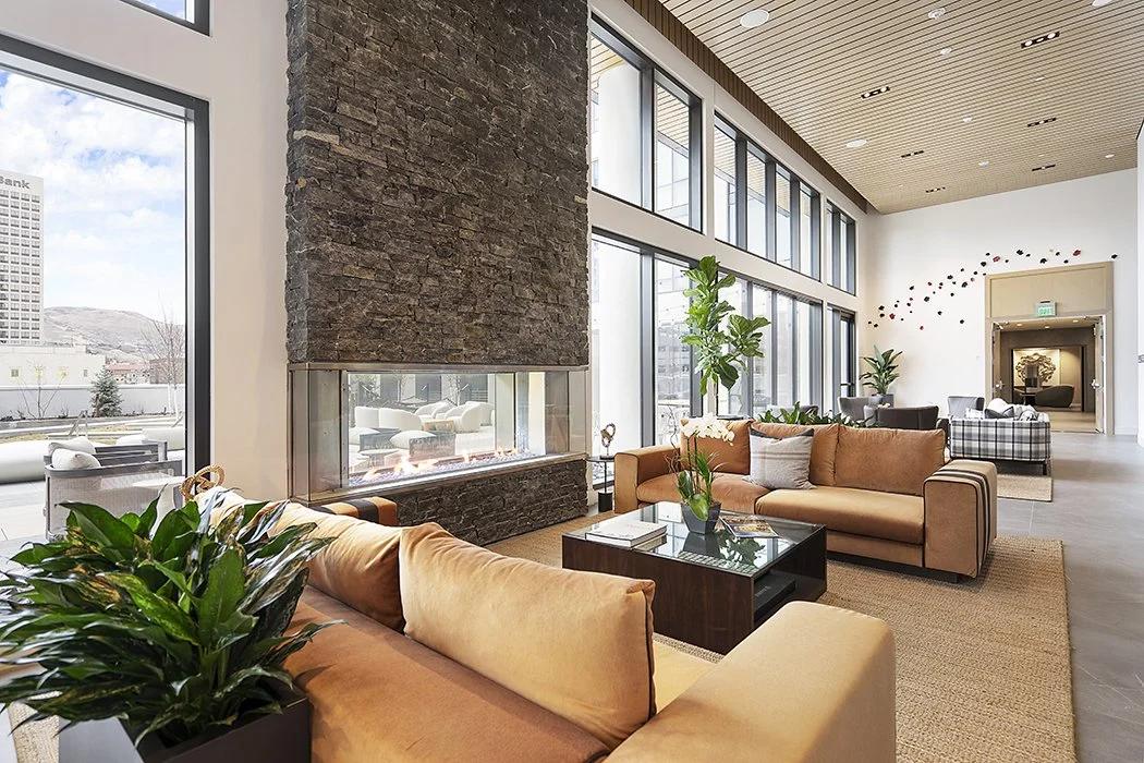 Modern hotel lobby with large floor-to-ceiling windows, beige sofas, potted plants, a glass fireplace, and a high ceiling with wood paneling.