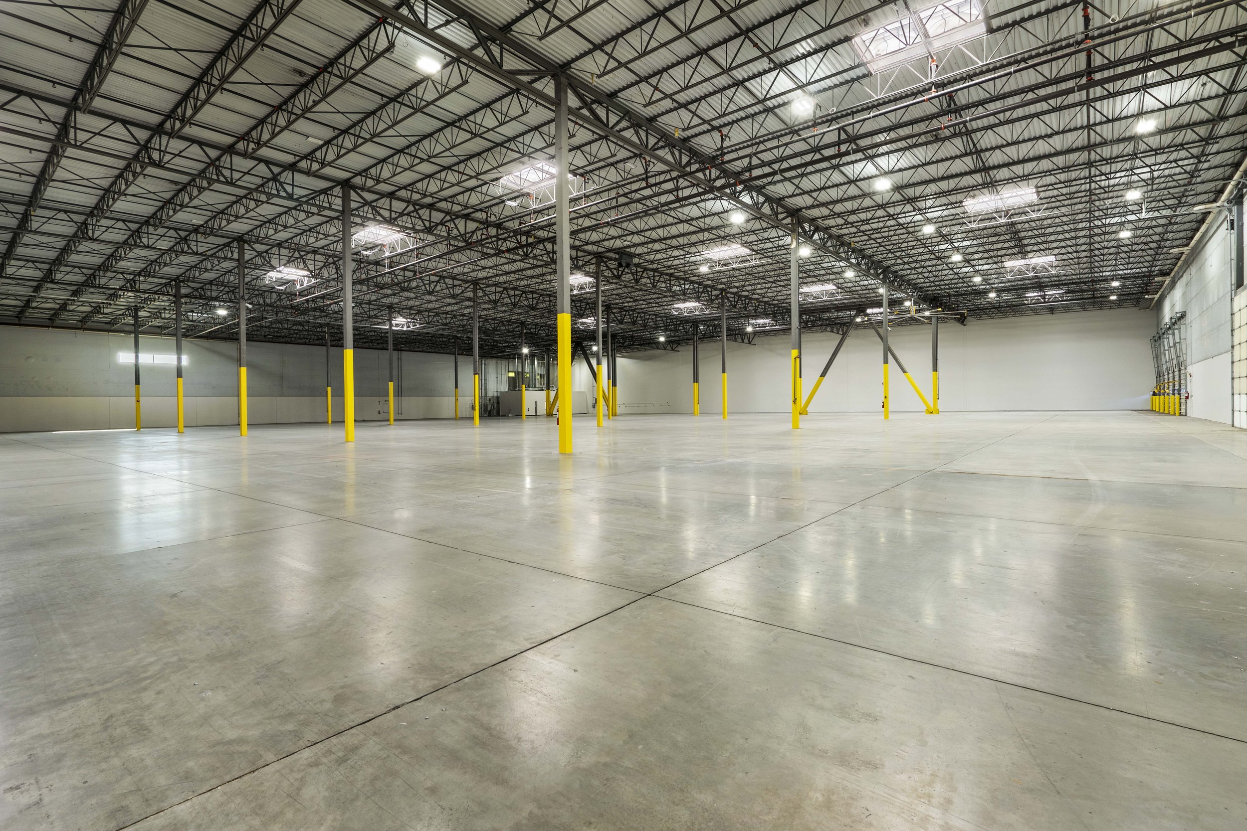 Empty warehouse with concrete floor, yellow safety poles, and high metal ceiling with bright overhead lighting.