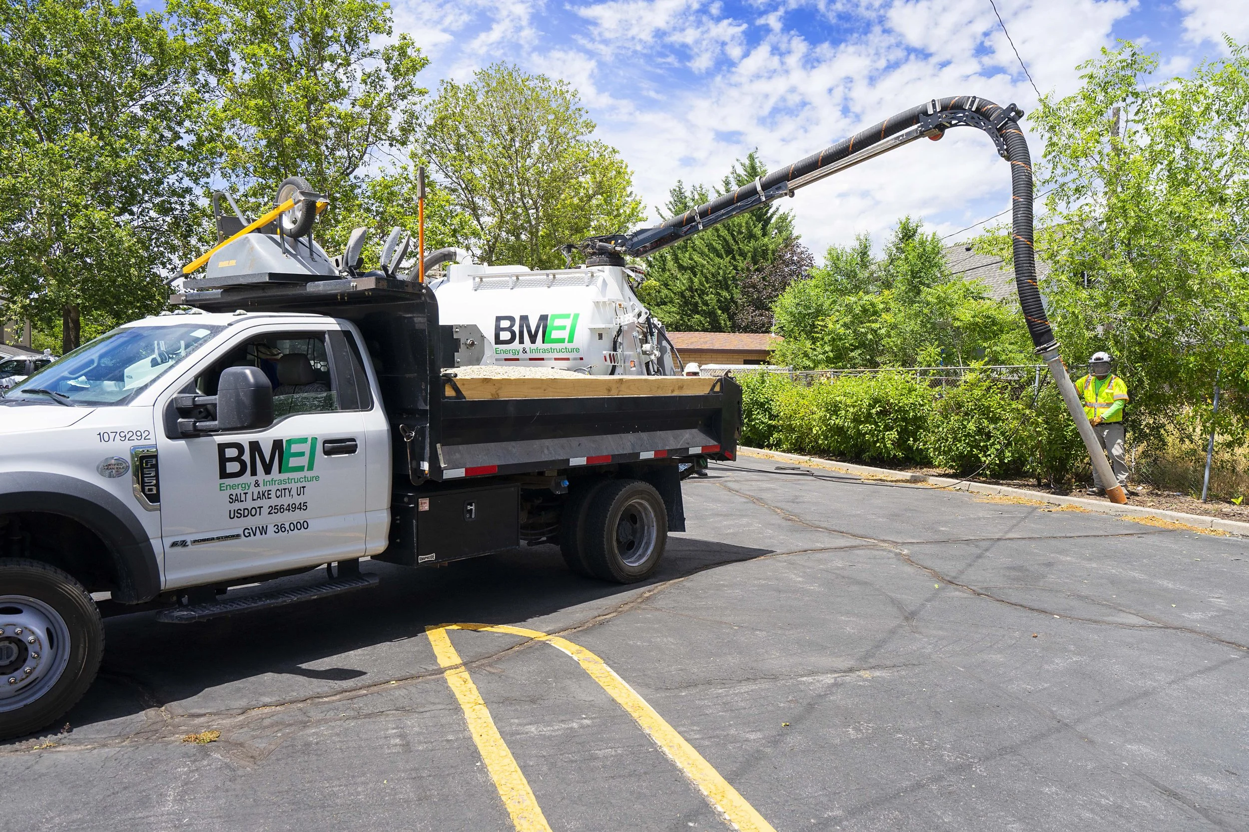 A utility truck with BMEI Energy and Infrastructure branding is extracting a tree stump from a parking lot using a large hose and equipment, with a worker nearby.