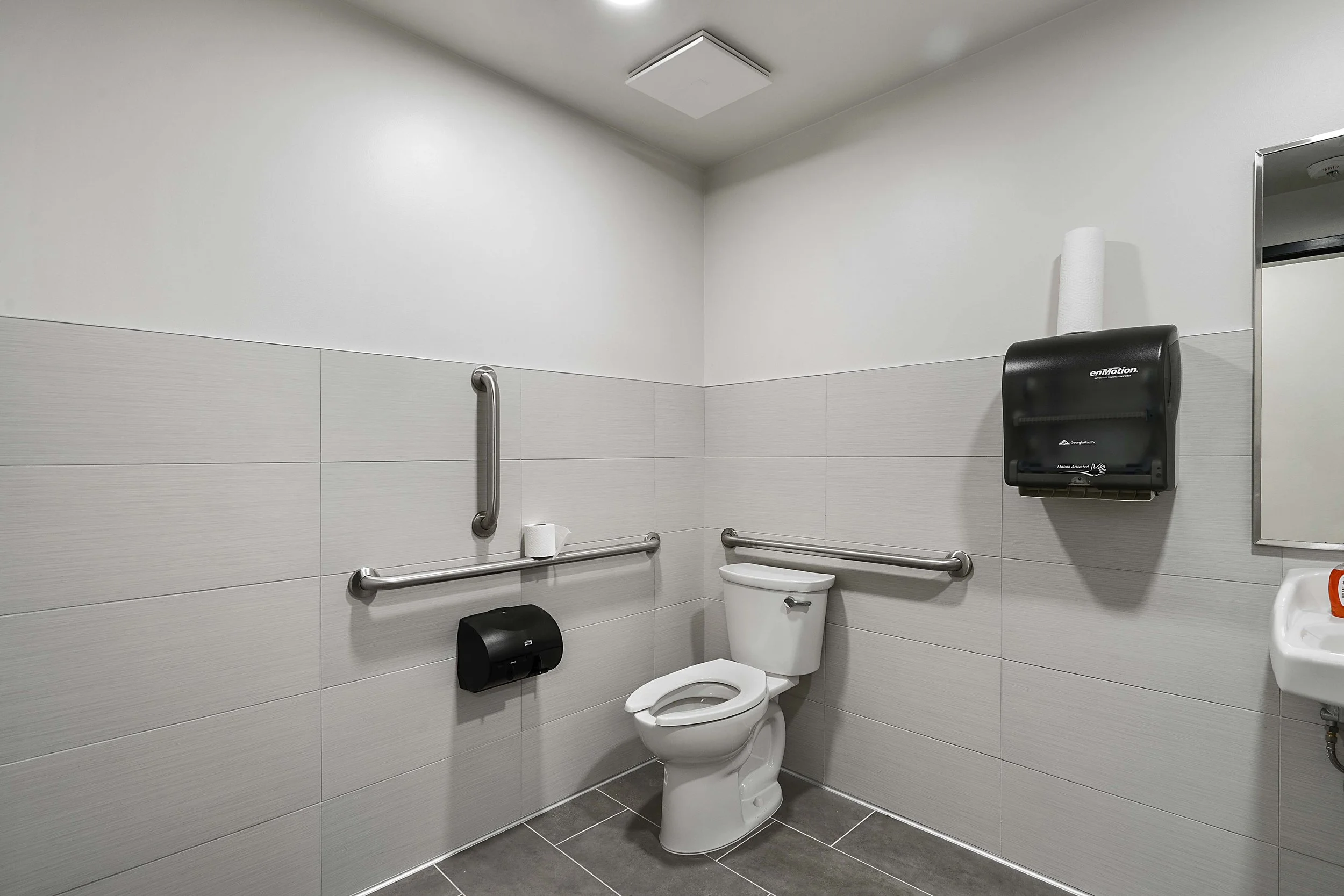 Accessible hospital bathroom with a toilet, grab bars, a wall-mounted paper towel dispenser, a mirror, and a small sink.