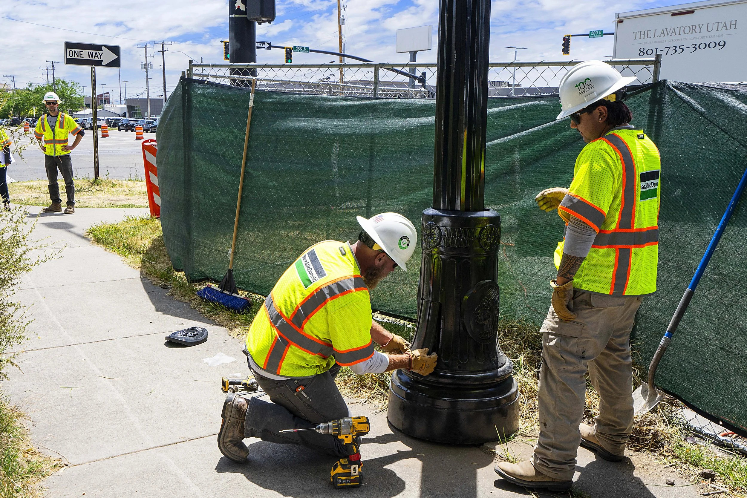 Two construction workers installing a street lamp at a sidewalk, wearing safety vests and helmets, with a person nearby on the sidewalk and a construction site fence in the background.