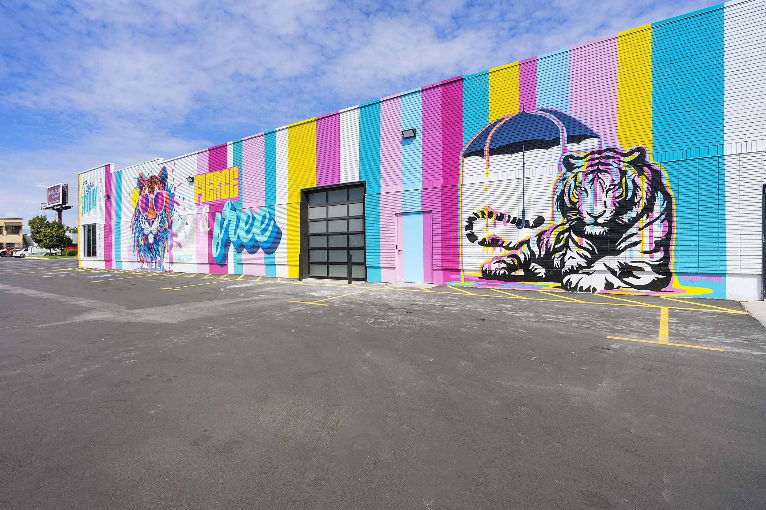 Colorful mural on the side of a building featuring a tiger with sunglasses, the words 'FIERCE and free,' and a black and white tiger with a rainbow-colored umbrella, with a parking lot in the foreground and a blue sky above.