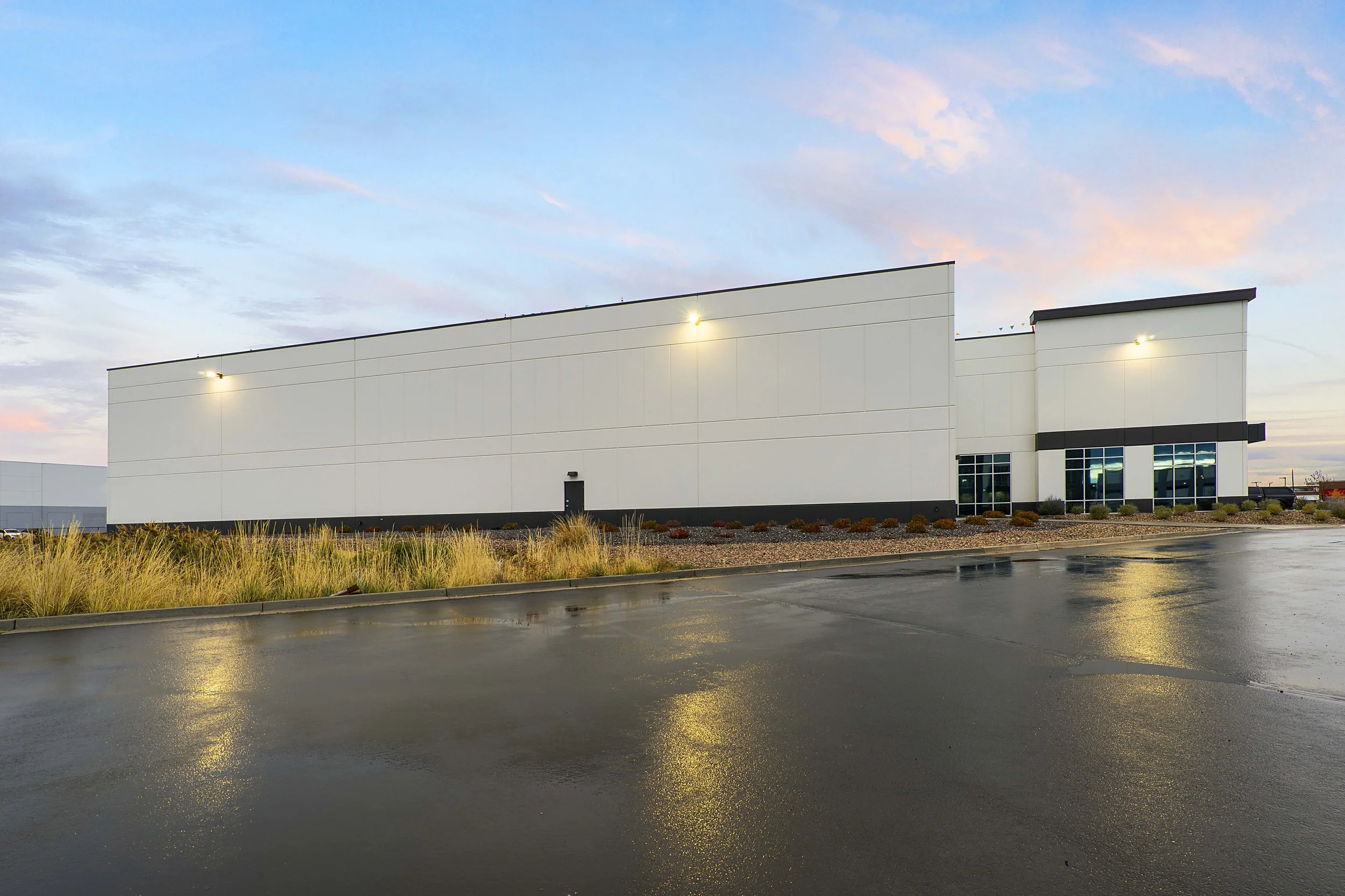 Empty commercial building with a white facade, large windows, and exterior lights, surrounded by a wet parking lot and landscaping under a pastel sky.