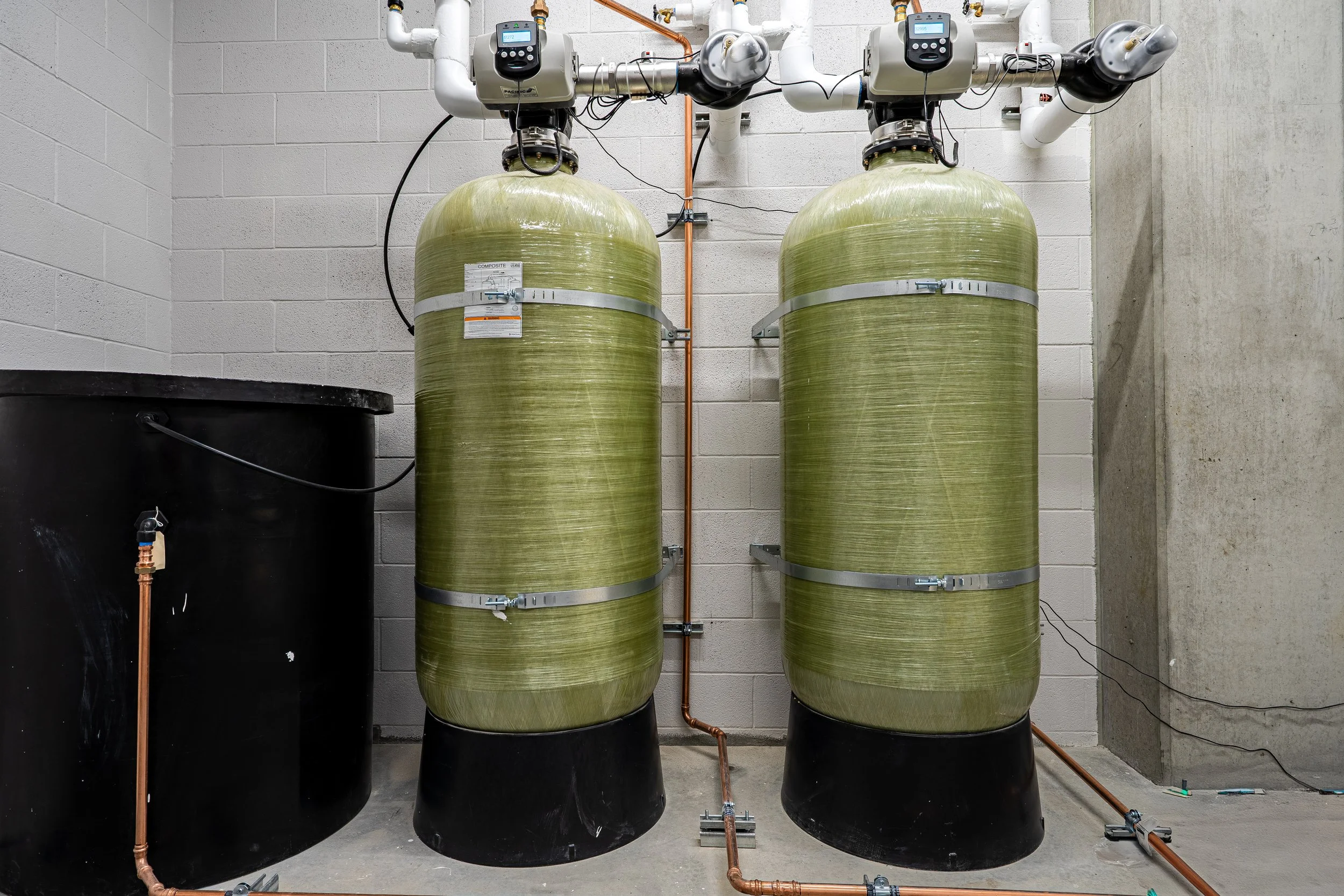 Two large green water filtration tanks with pipes and controls on a concrete wall in an industrial setting.