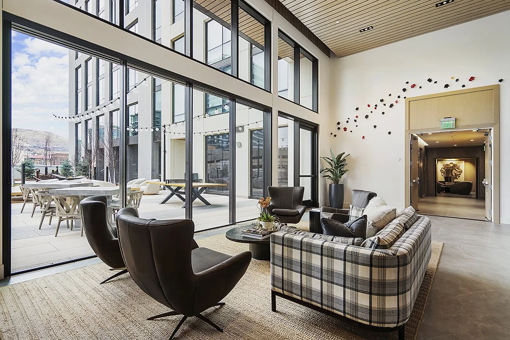 Modern lobby with large glass windows, black chairs, plaid sofa, potted plant, and outdoor patio with tables and chairs.