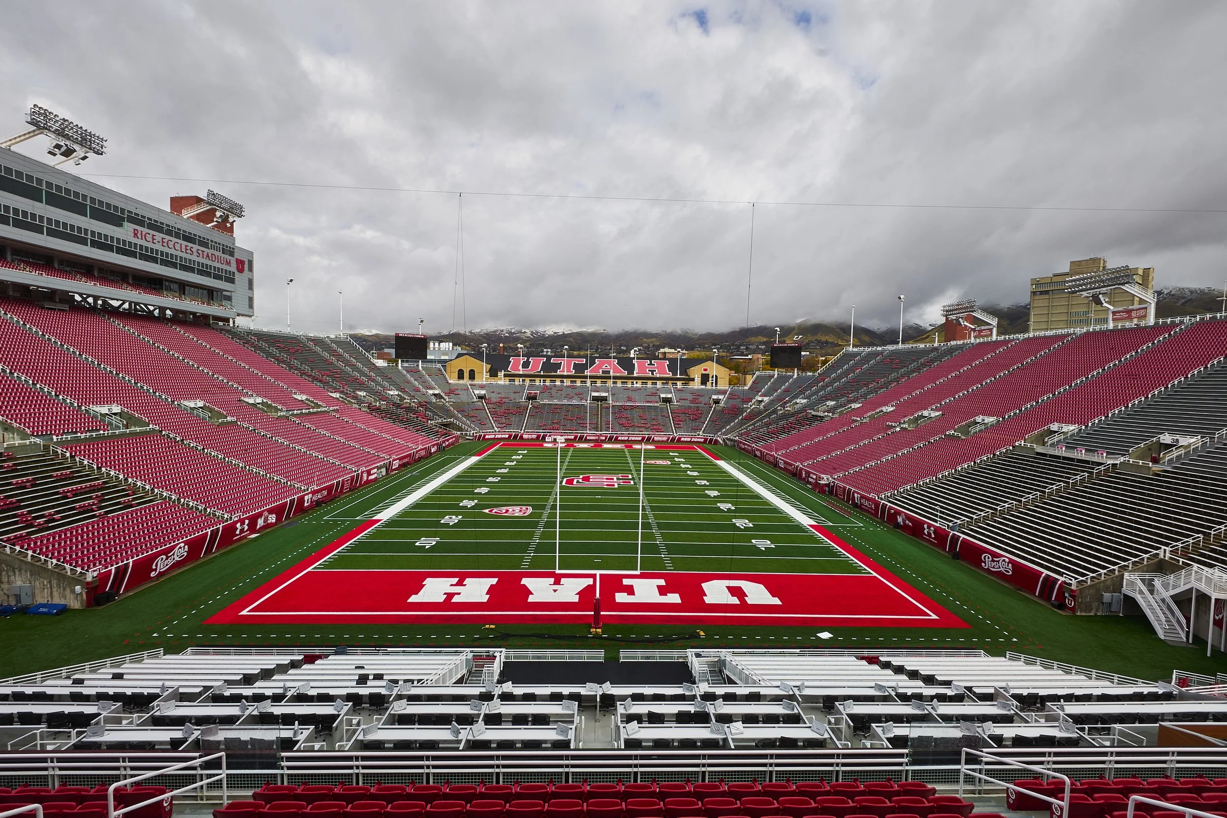 Empty football stadium with red seats, green field, and cloudy sky, featuring the university name Utah displayed in the background.