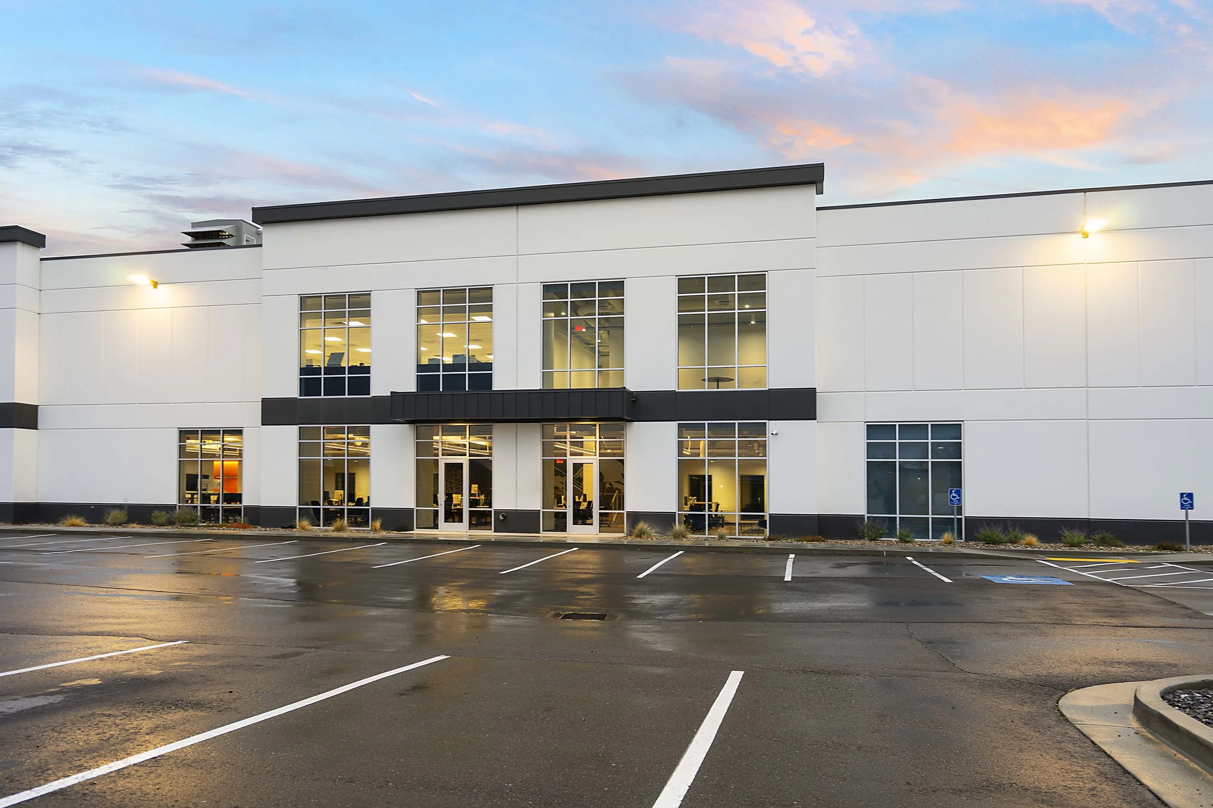 Modern office building with large glass windows and an empty parking lot in front, taken at dusk with a colorful sky.