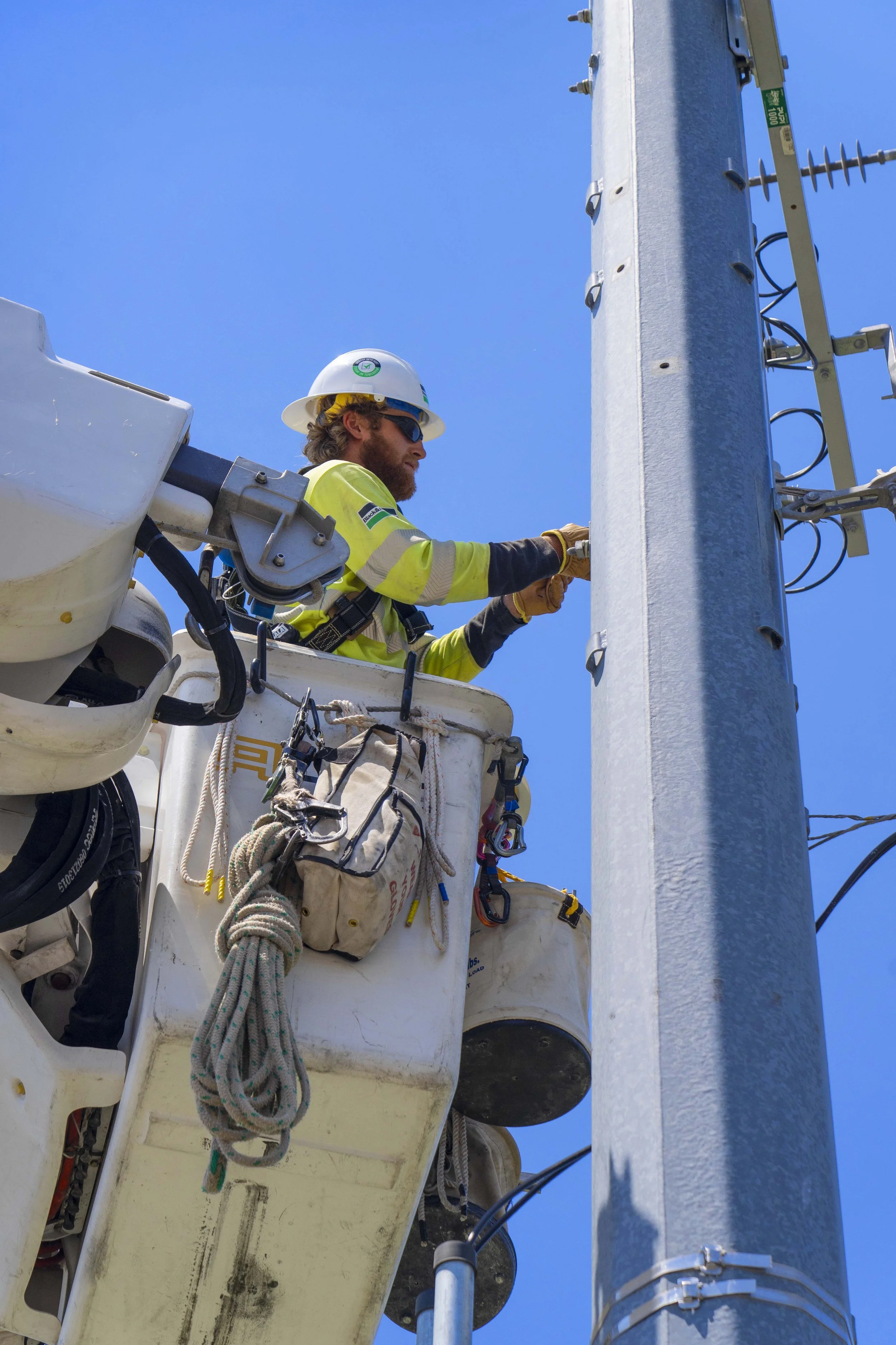 A utility worker wearing a hard hat, sunglasses, and a safety vest is working on power lines from a bucket lift against a clear blue sky.