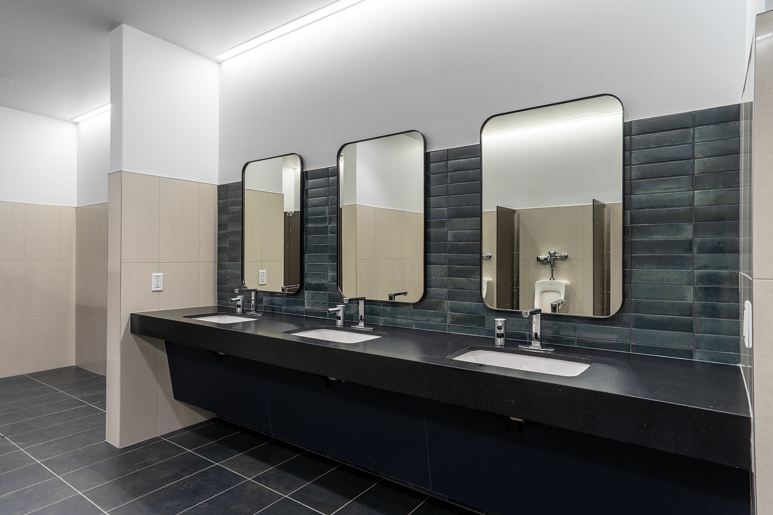 Public men's restroom with three sinks, mirrors, black countertop, tan and green tiled walls, and urinals reflected in the mirrors.