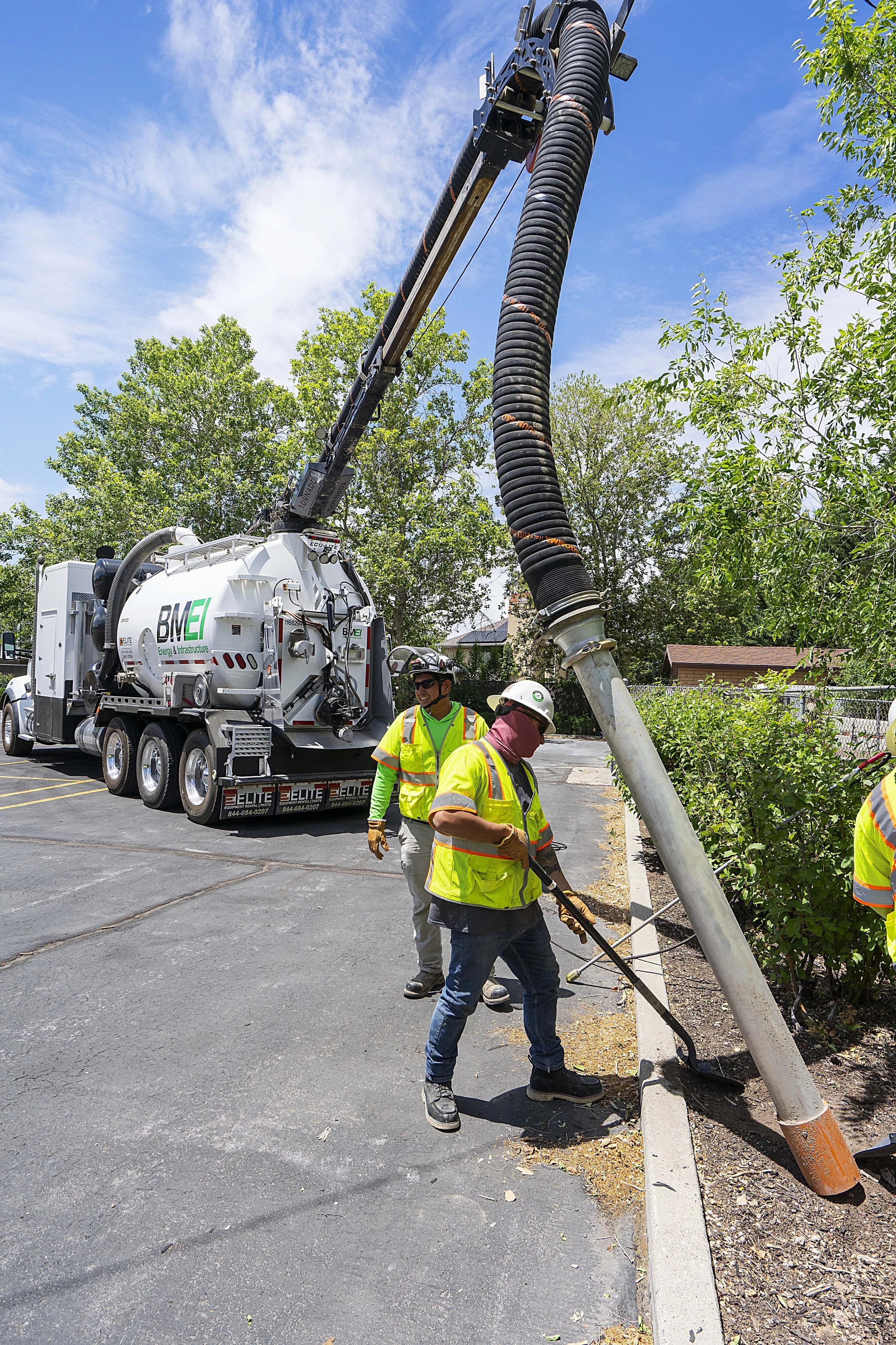 Workers in safety gear repair or maintain a fallen street light pole near a parking lot, with a truck and greenery in the background.