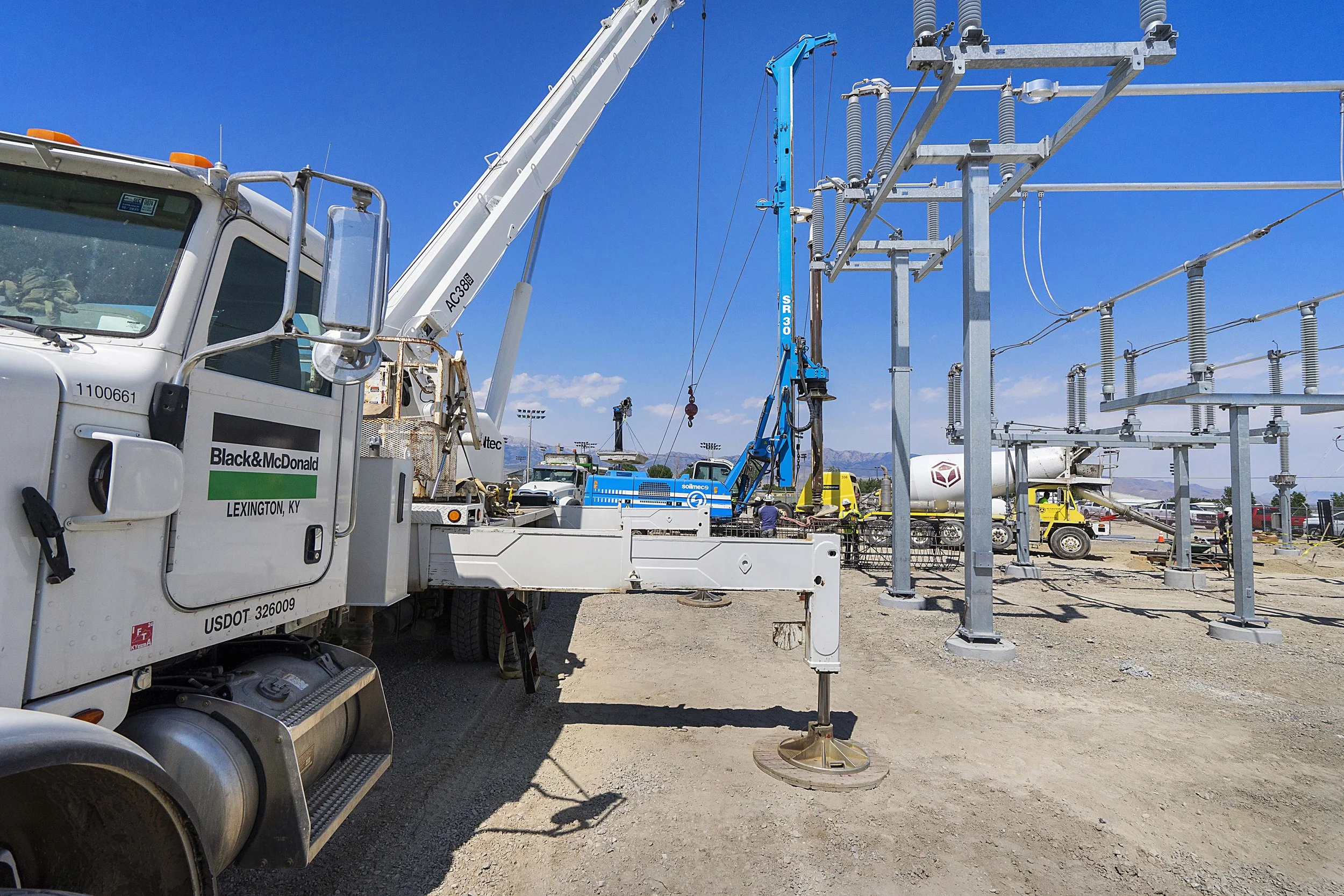 Construction workers using cranes and equipment to build an electrical substation with power lines and transformers under a clear blue sky.