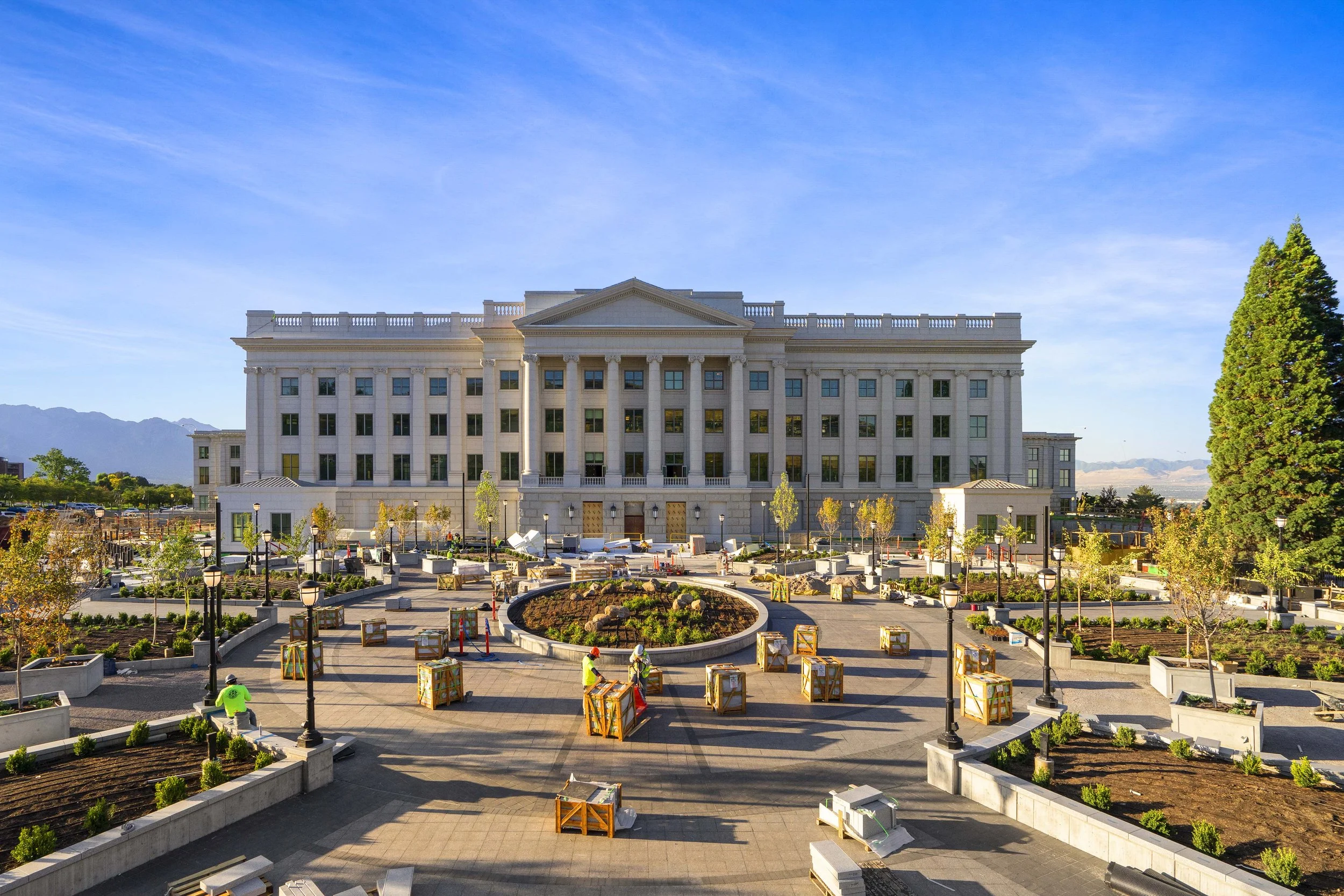 Large white government or institutional building under construction, with workers setting up the area in a landscaped plaza with trees, benches, and lampposts, mountains in the background.