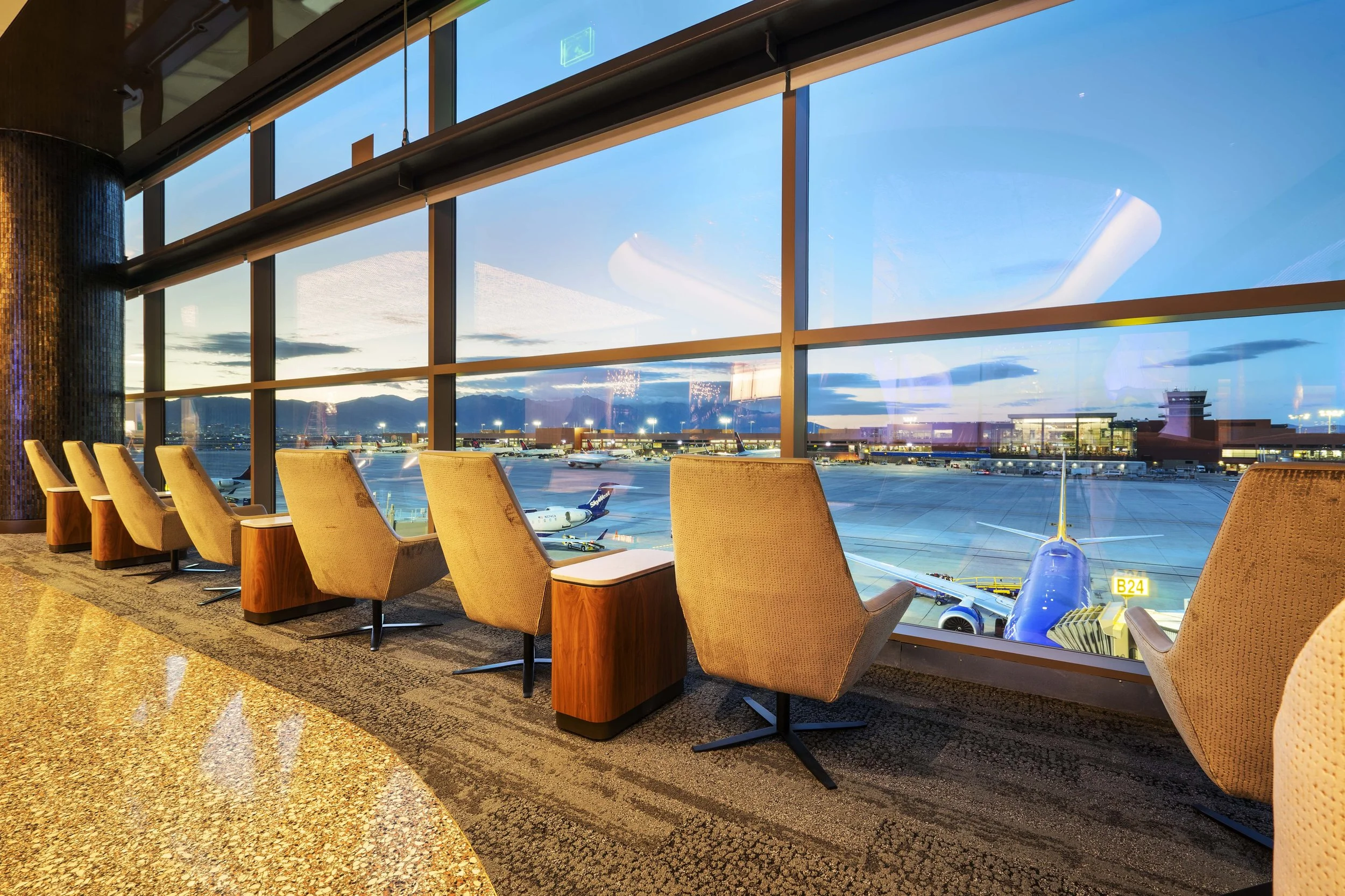 Airport lounge with beige chairs facing large windows overlooking the runway and parked airplanes at dusk.