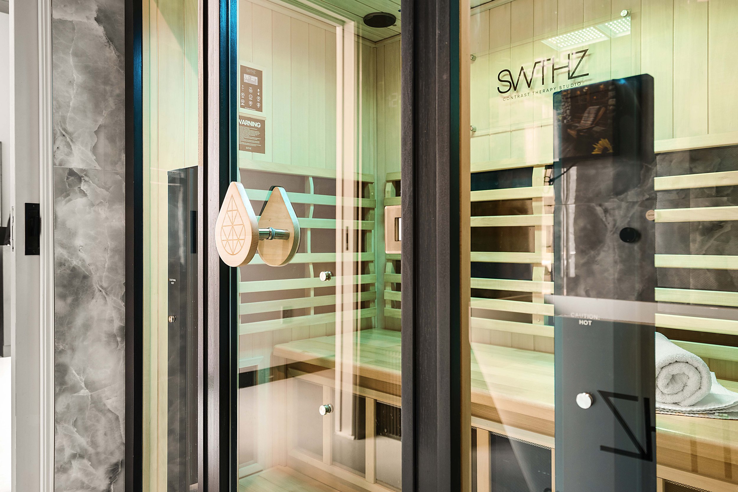 A wooden infrared sauna inside a glass enclosure with a towel on the bench and a sign that reads "SWTHZ Contrast Therapy Studio."