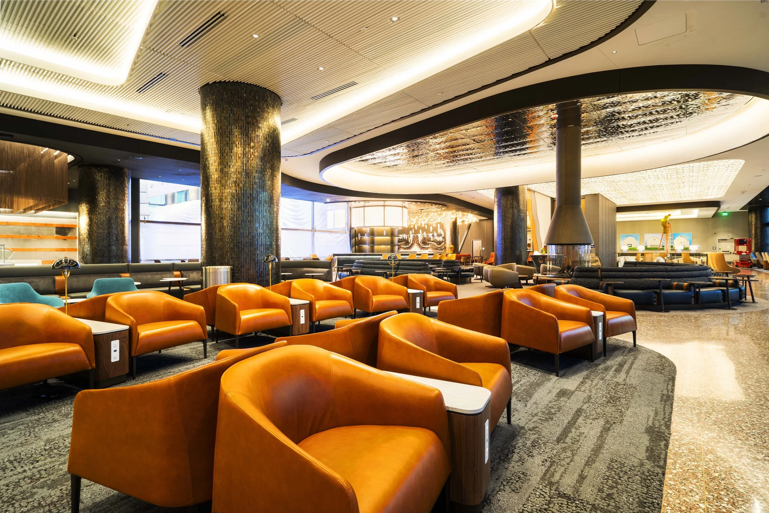 Modern airport lounge with orange leather armchairs, black pillars, a fireplace, and a play area in the background.