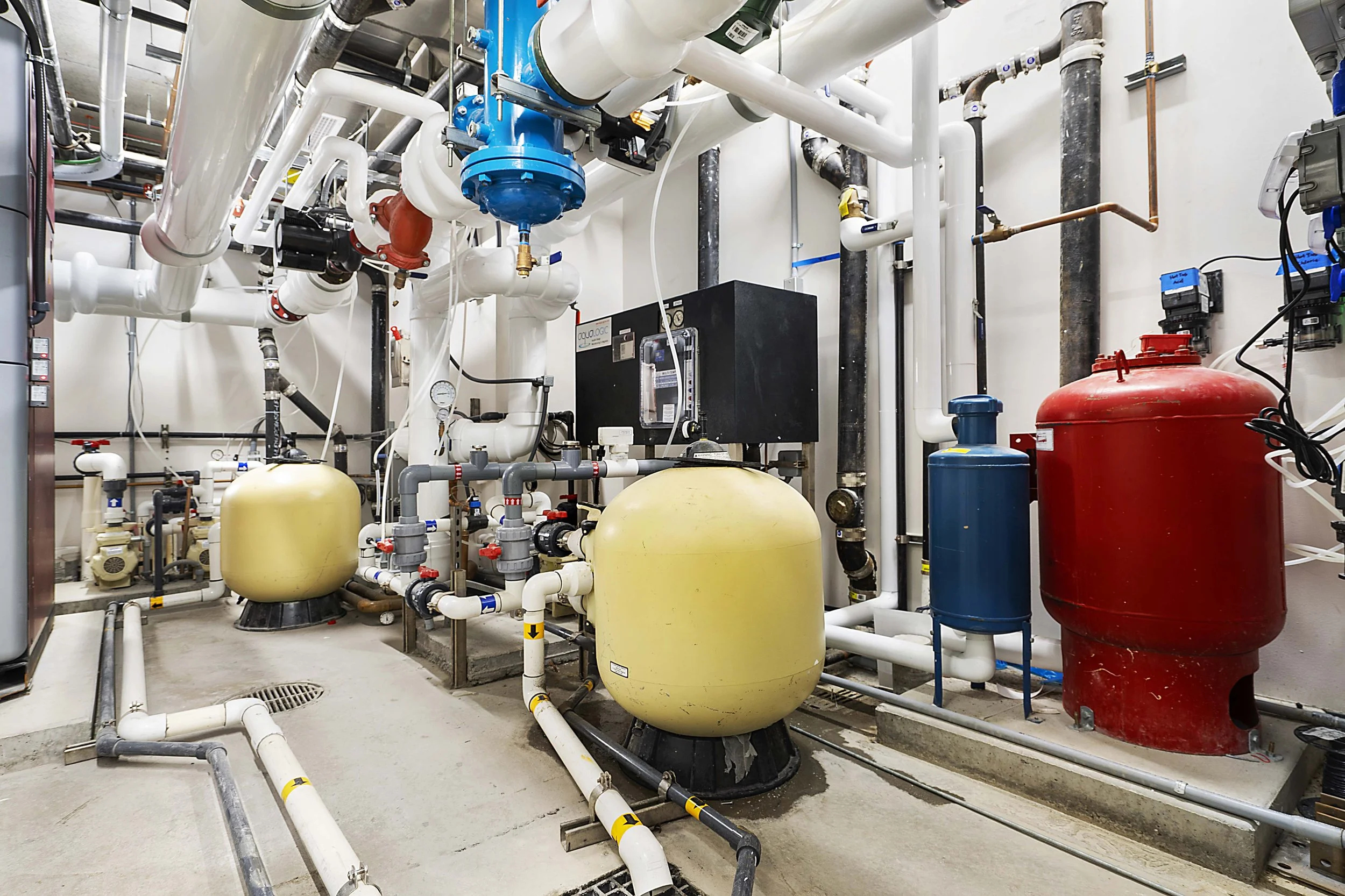 Mechanical room with various pipes, tanks, and equipment for water or HVAC systems, including a large red tank, yellow tanks, and several interconnected pipes and control devices.