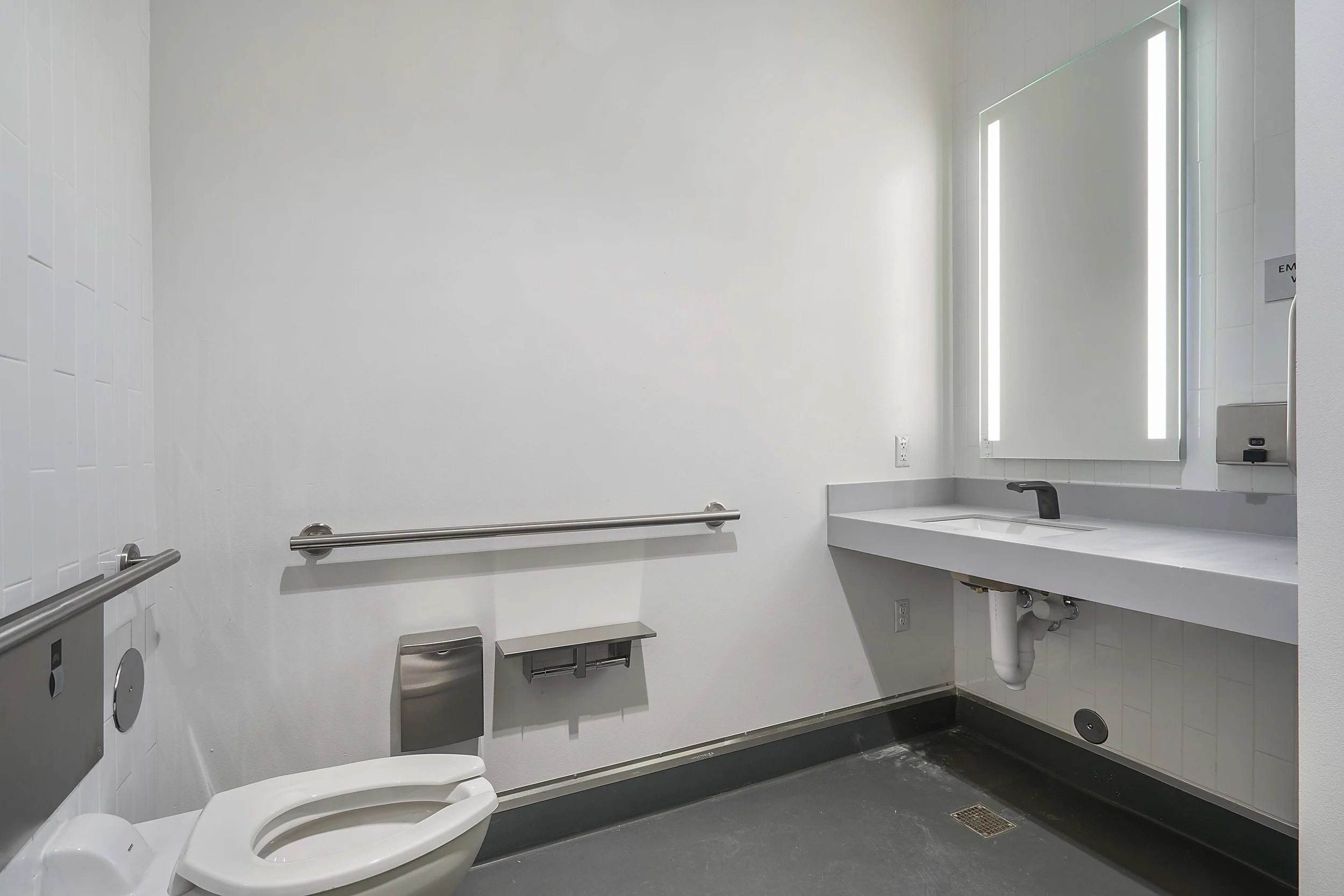 Accessible bathroom with white walls, a toilet, grab bars, a sink with a large mirror, and an emergency call button.