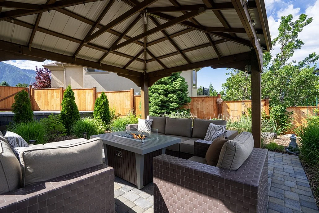 Outdoor patio with fire pit under a gazebo, surrounded by wicker furniture with cushions, set in a landscaped backyard with a wooden fence and trees.