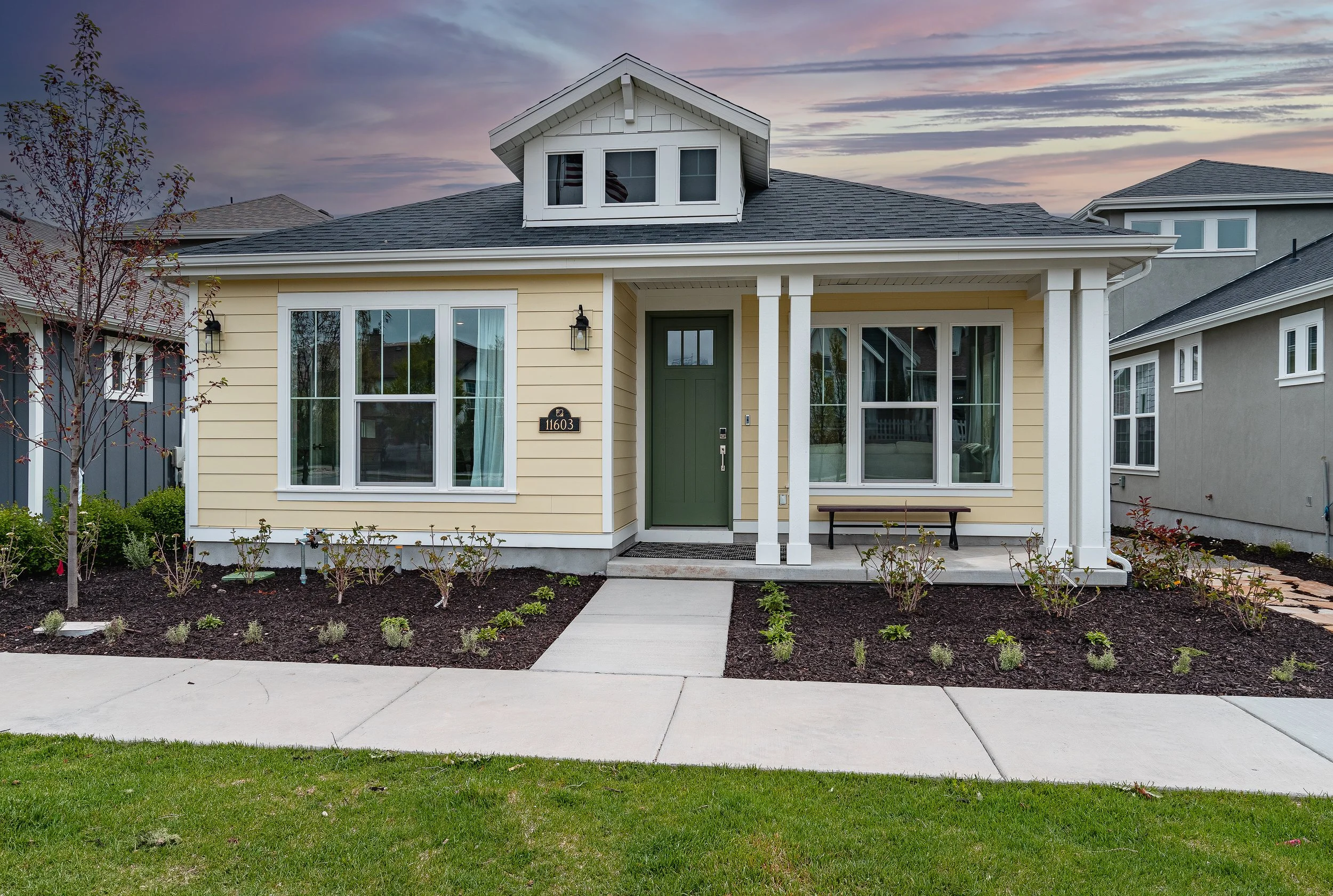 Single-story yellow house with green door, front porch, and landscaped yard at sunset.