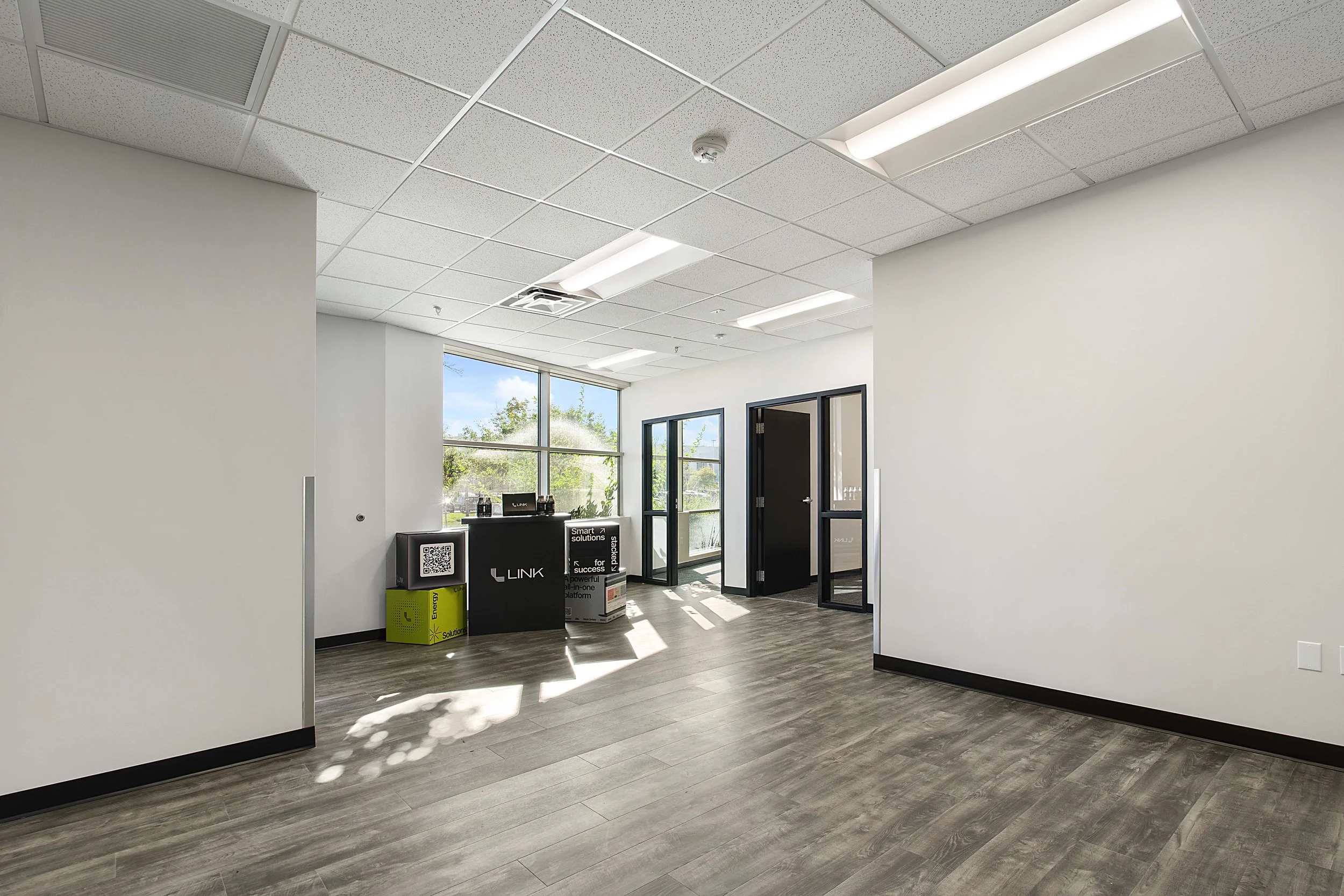Empty office space with large window, grey wood floor, white walls, and ceiling with fluorescent lights. Small counter with tech products near the window.