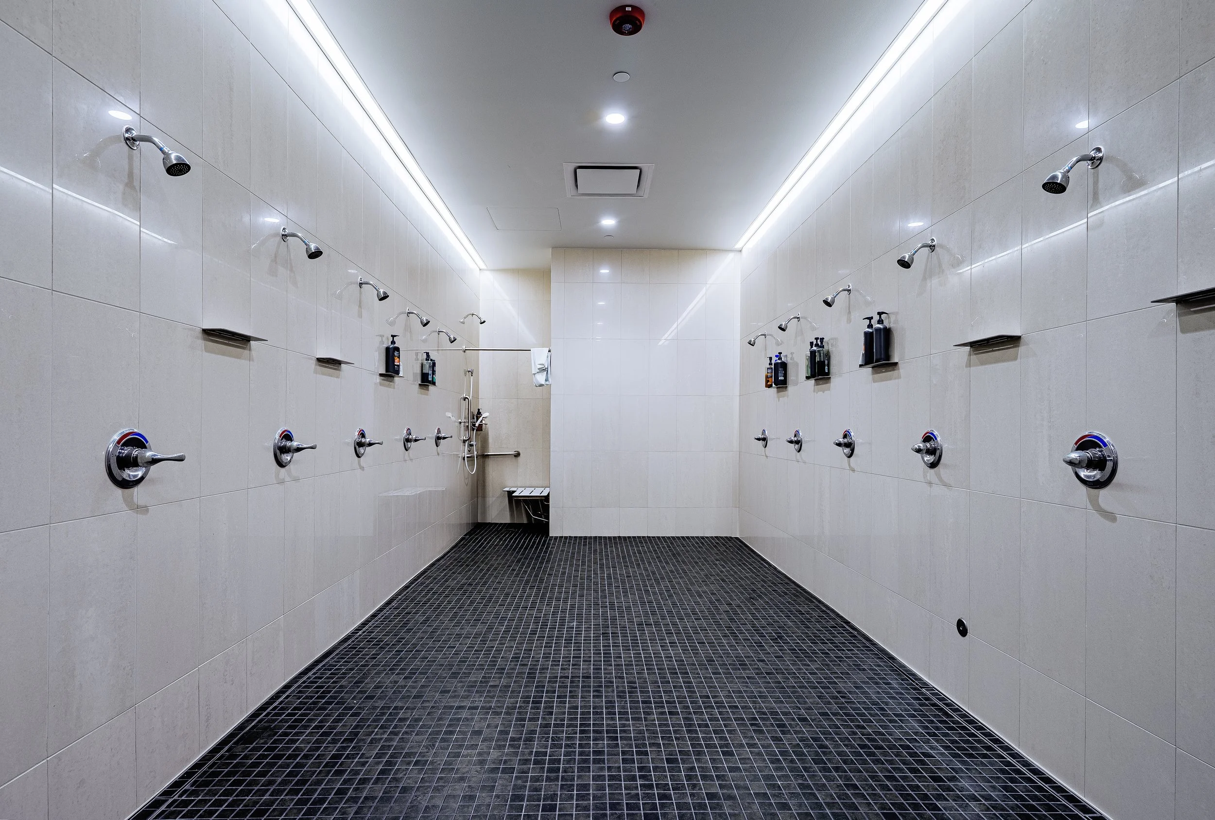 A modern communal shower room with multiple shower heads and bottles of shampoo and soap on small shelves, tiled walls, black tiled floor, and a ceiling with LED lighting.