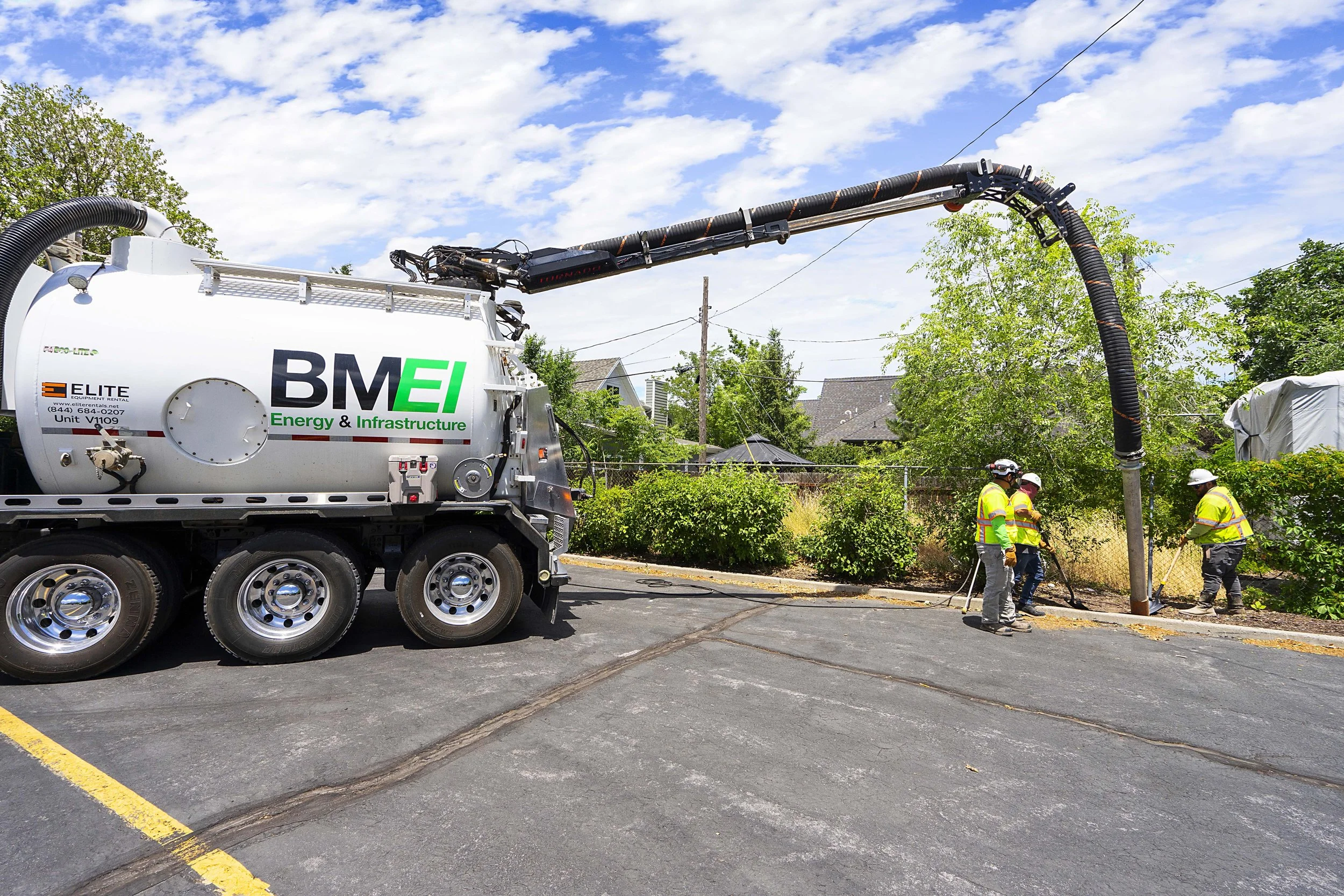 Workers in safety vests and helmets operate equipment related to underground utility installation or repair on a residential street, with a large vacuum truck in the background and trees lining the street.