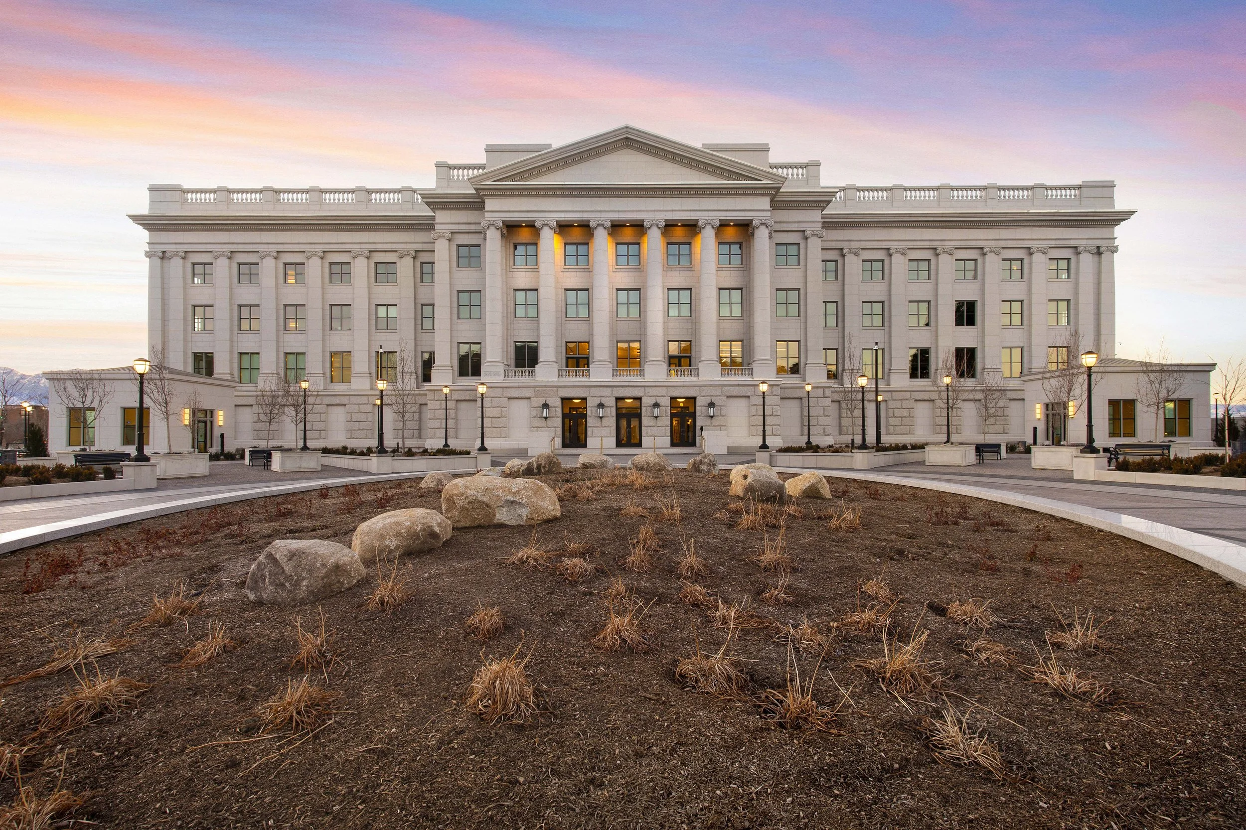 Front view of a large, white neoclassical style government building at sunset, with a landscaped foreground, lampposts lining the walkway, and a colorful sky.