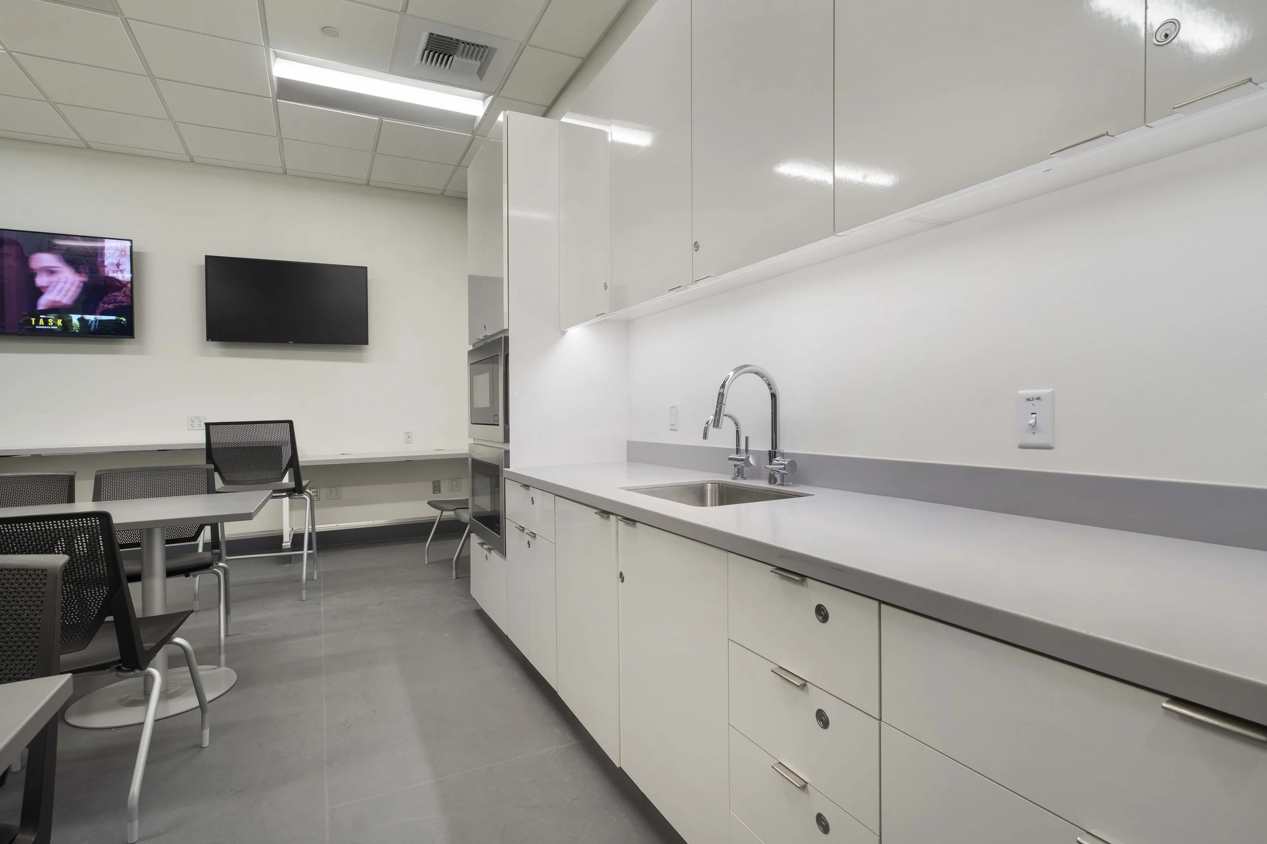Empty break room with white cabinets, a sink, a microwave, and two televisions on the wall.