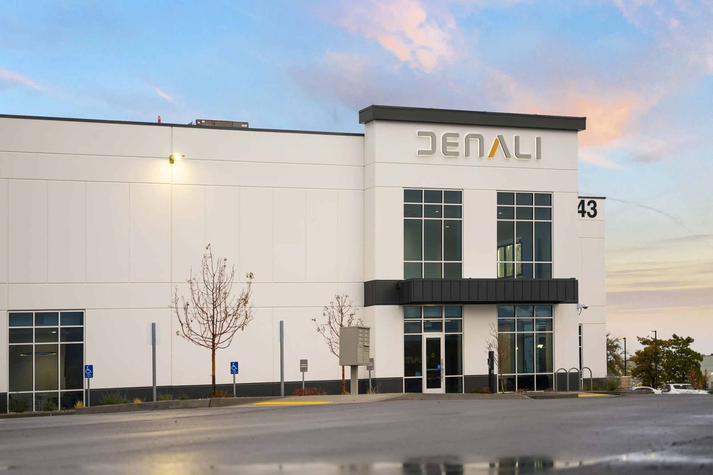 A modern commercial building with a white facade and large glass windows, displaying the number 43, situated in a parking lot with a few small trees and handicap parking signs, during dusk with a colorful sky.