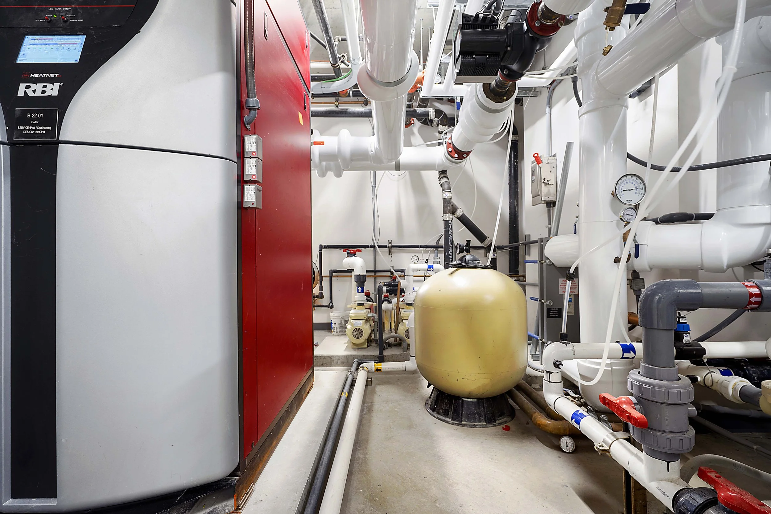 A mechanical room with various pipes, gauges, and equipment including a large yellow tank, a red control panel, and a white and gray boiler or water heater.