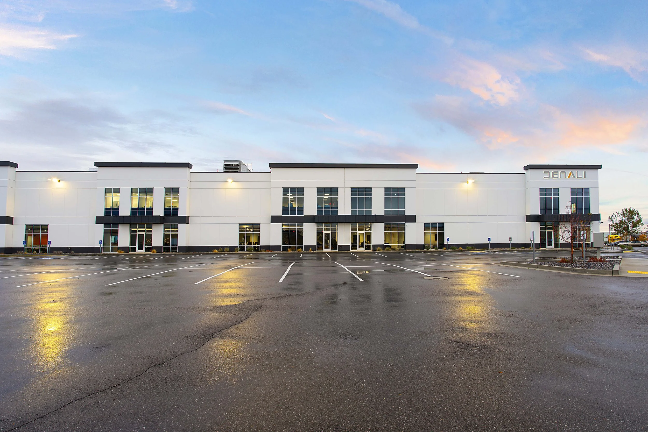 Empty parking lot in front of a modern white commercial building with multiple windows and a sign that reads 'DENA.' The sky is partly cloudy with pink and blue hues.