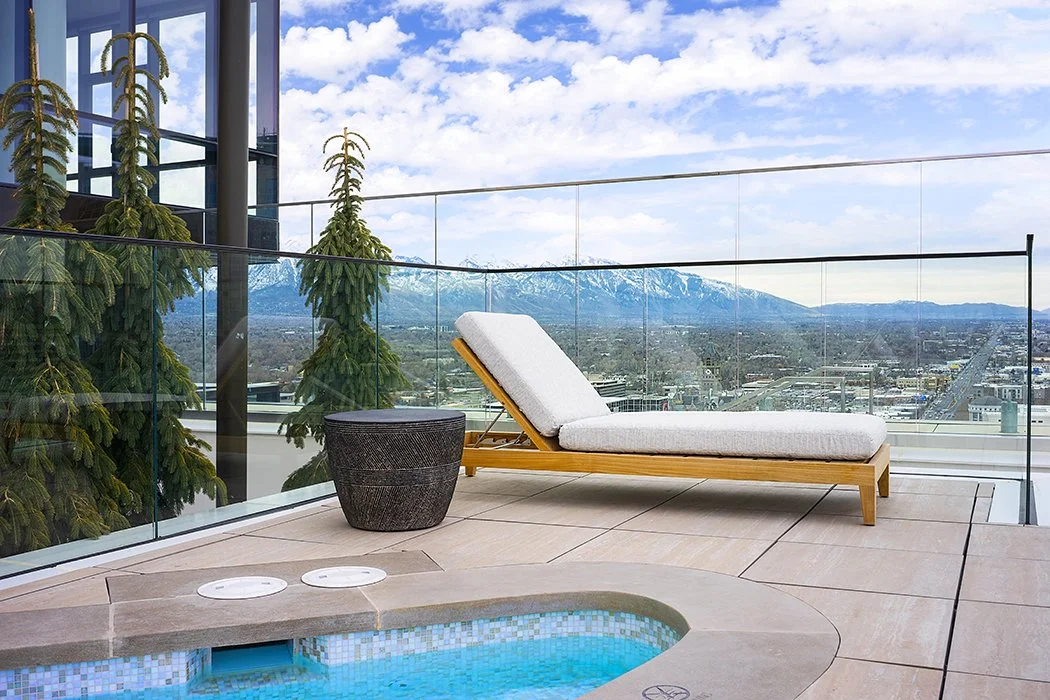 A rooftop balcony with a white lounge chair, a black side table, and a small pool overlooking a cityscape with mountains in the background.