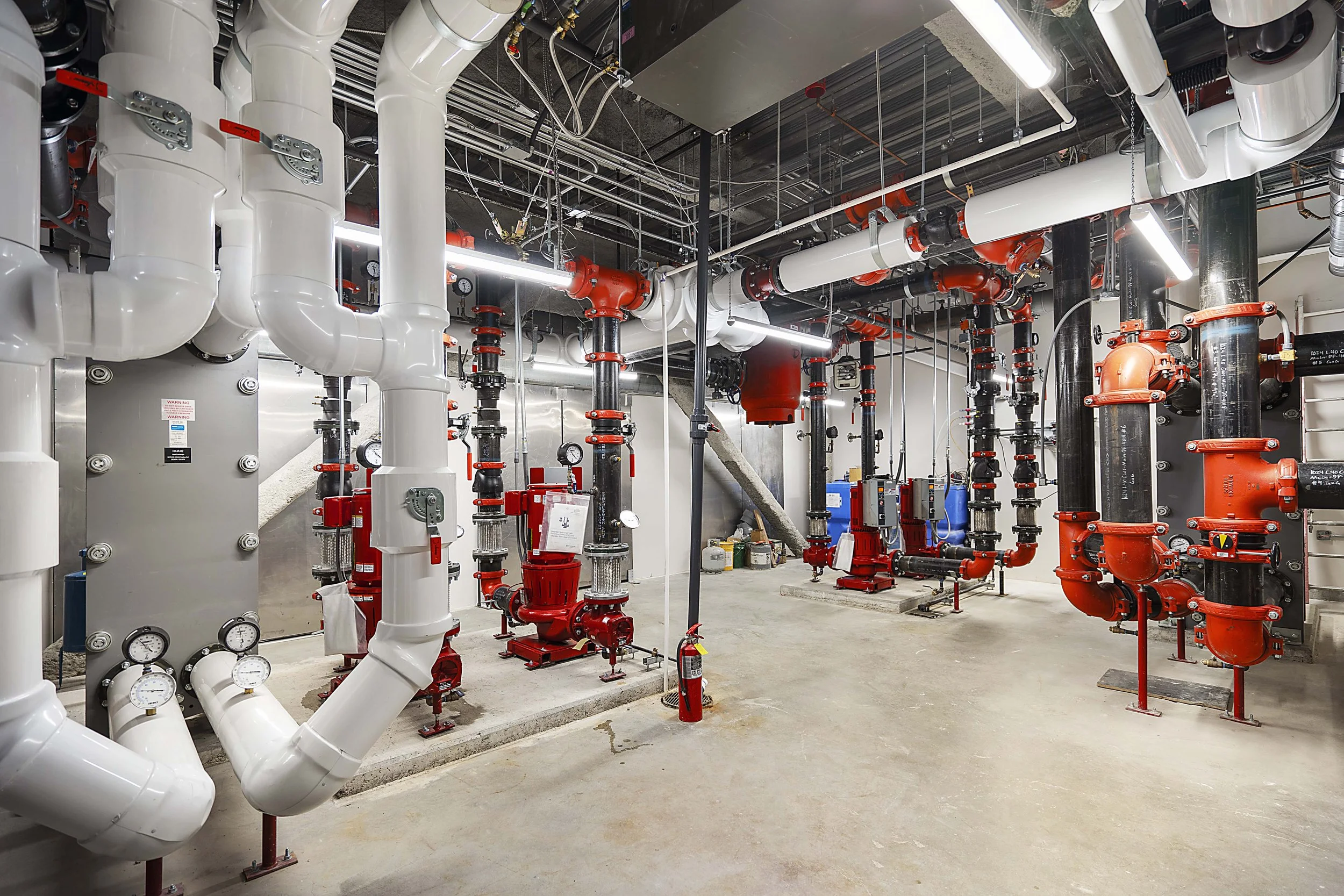Industrial mechanical room with white and black pipes, gauges, valves, and equipment, illuminated by overhead fluorescent lights.