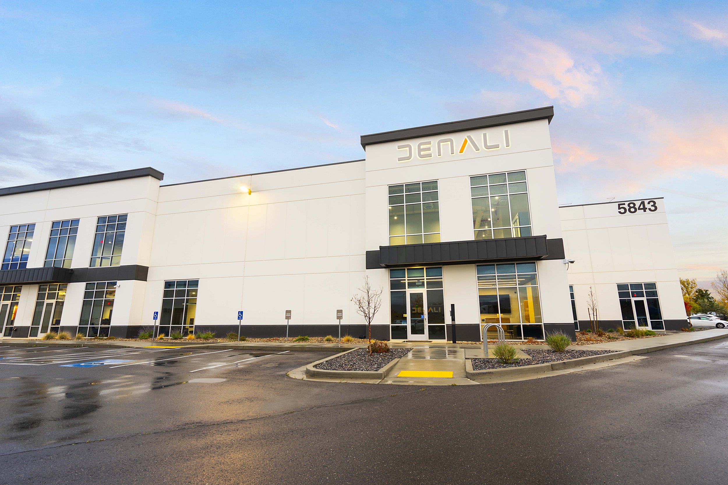 Exterior of a modern white commercial building with large windows, a black awning, and the name 'JENALI' on the upper section. Parking lot in front with a few handicap spaces and a small landscaped area.
