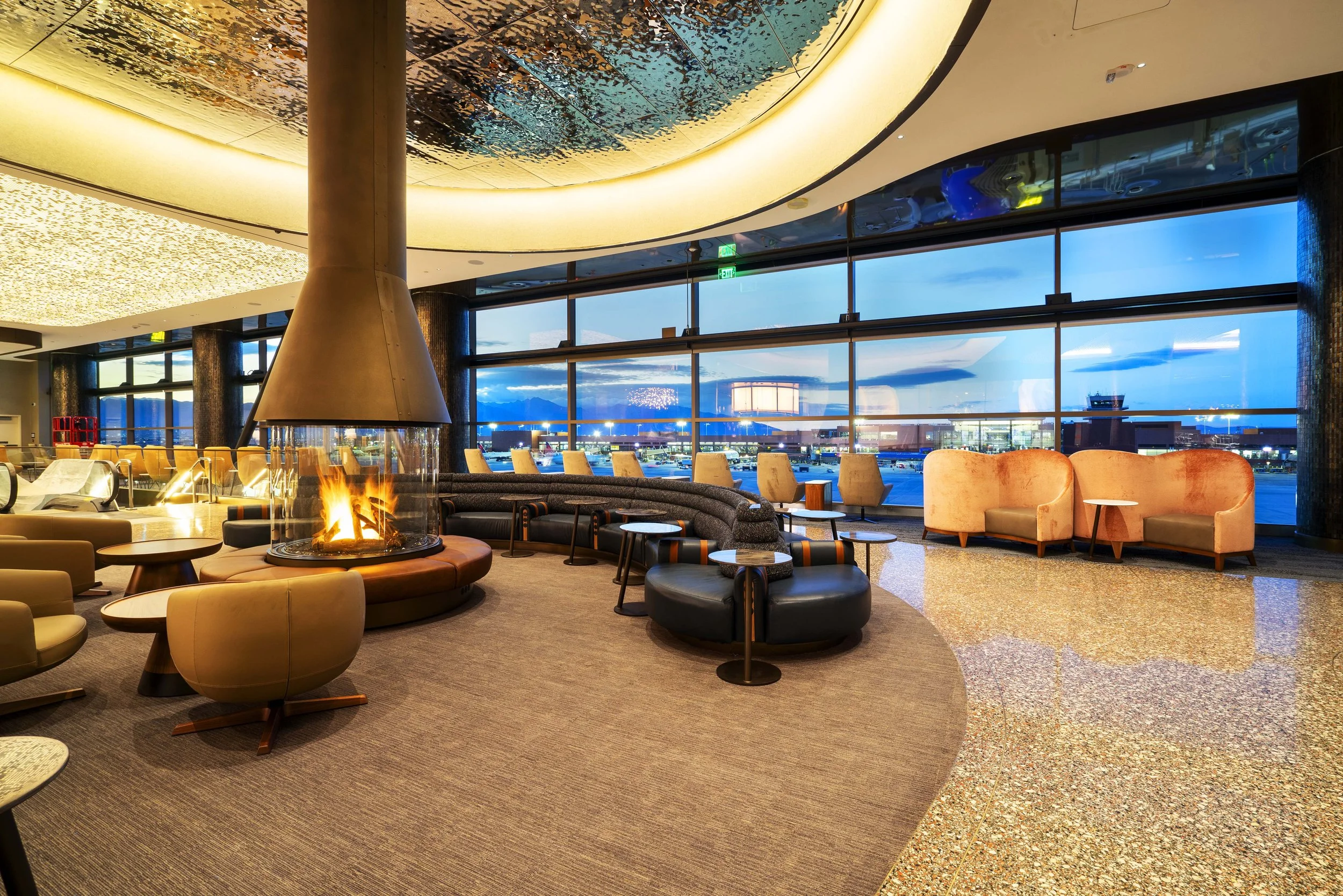 Airport lounge with large windows showing a blue evening sky, a central fireplace, and modern seating including armchairs and sofas.