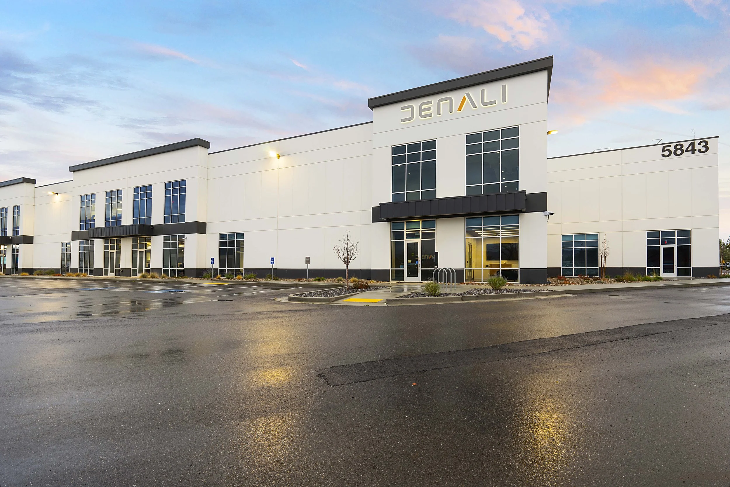 Modern white commercial building with black accents and large glass windows, identified as DENALI, located at 5843, with an empty parking lot and a few small trees in the landscape, during an overcast evening.
