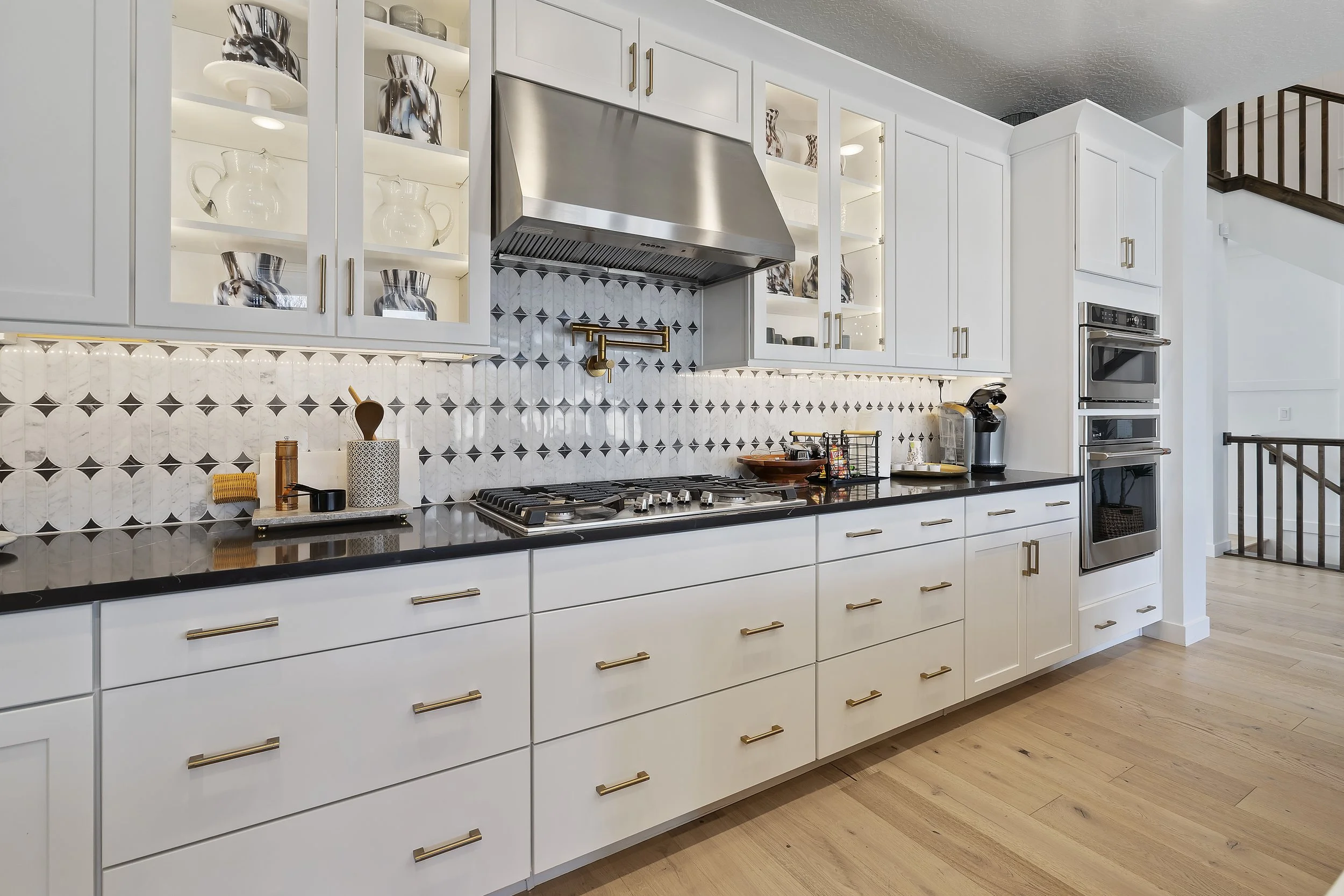 Modern kitchen with white cabinets, black countertops, and a geometric tile backsplash. Stainless steel range hood, built-in oven, and a countertop coffee maker are visible. Light wood flooring and open shelving with decorative items can also be seen