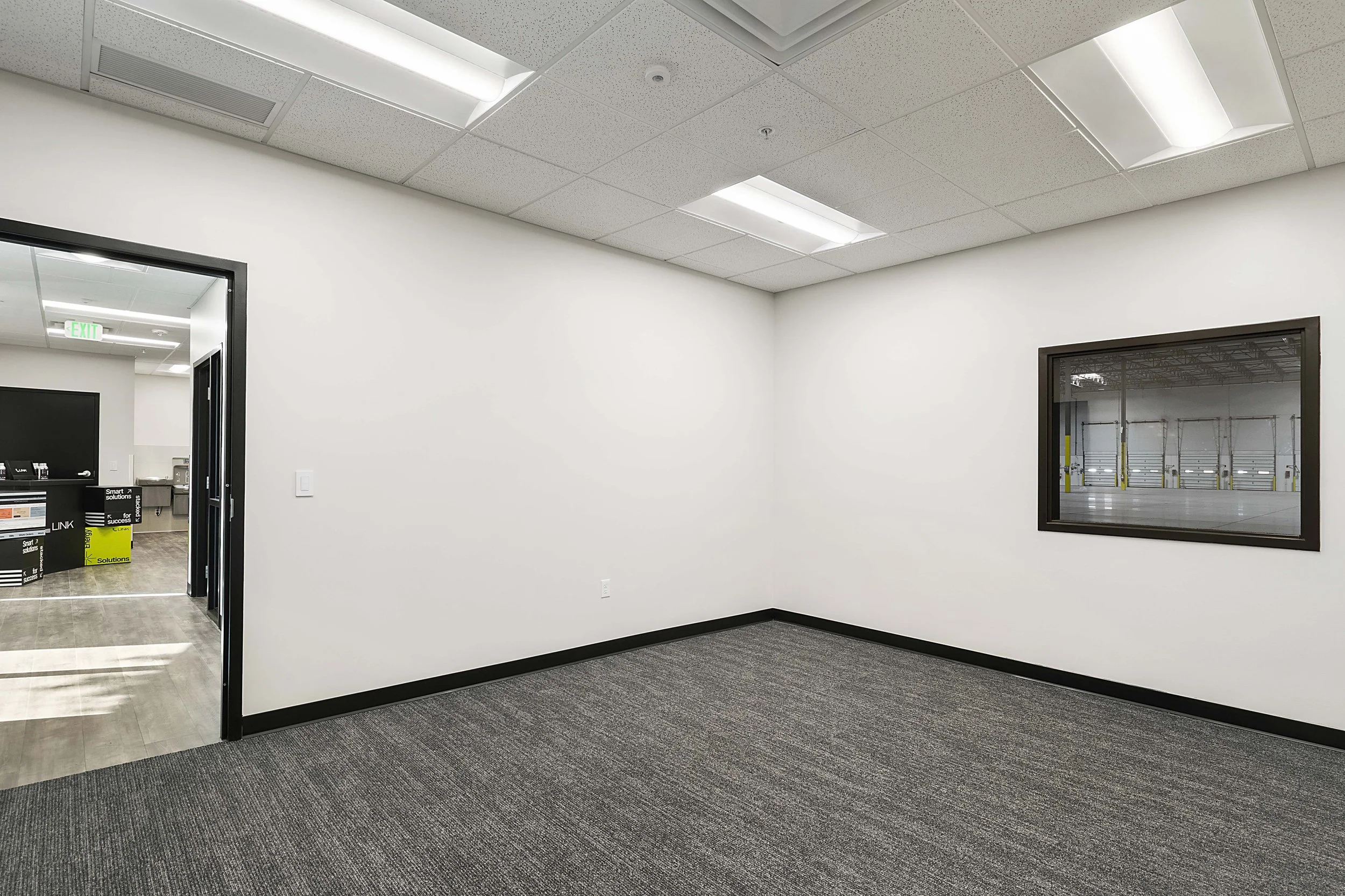 Empty office room with white walls, gray carpet, and a window looking into an empty warehouse or garage area.