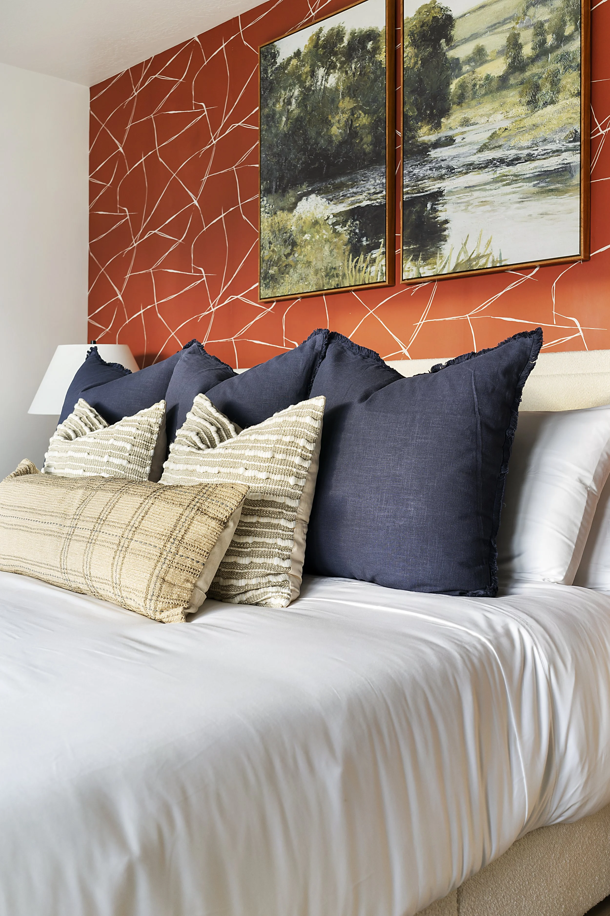 A bedroom with a bed featuring multiple pillows, artwork with trees and a river on a red accent wall, and a white lamp on the left side.