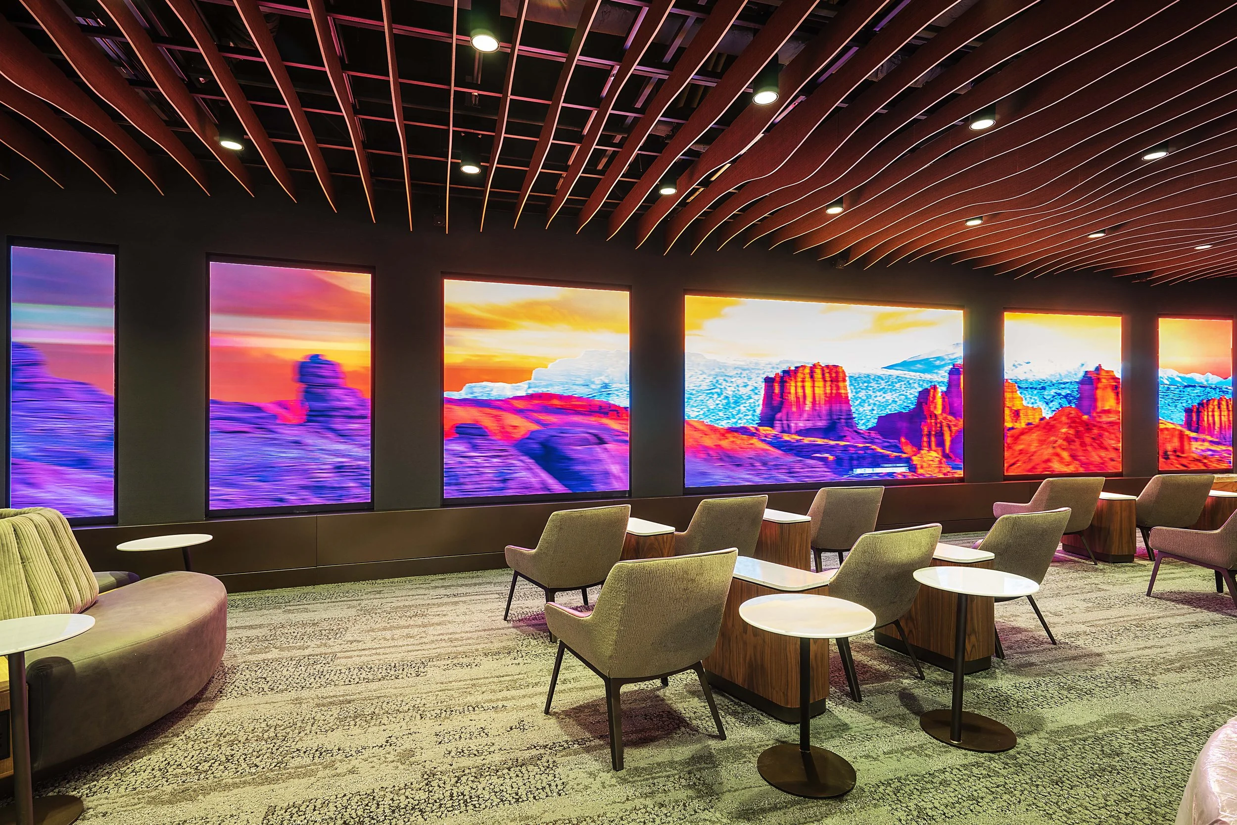 A modern lounge area with plush chairs and small white tables, large scenic windows displaying a vibrant desert landscape at sunset, with mountains and rock formations in bright colors.