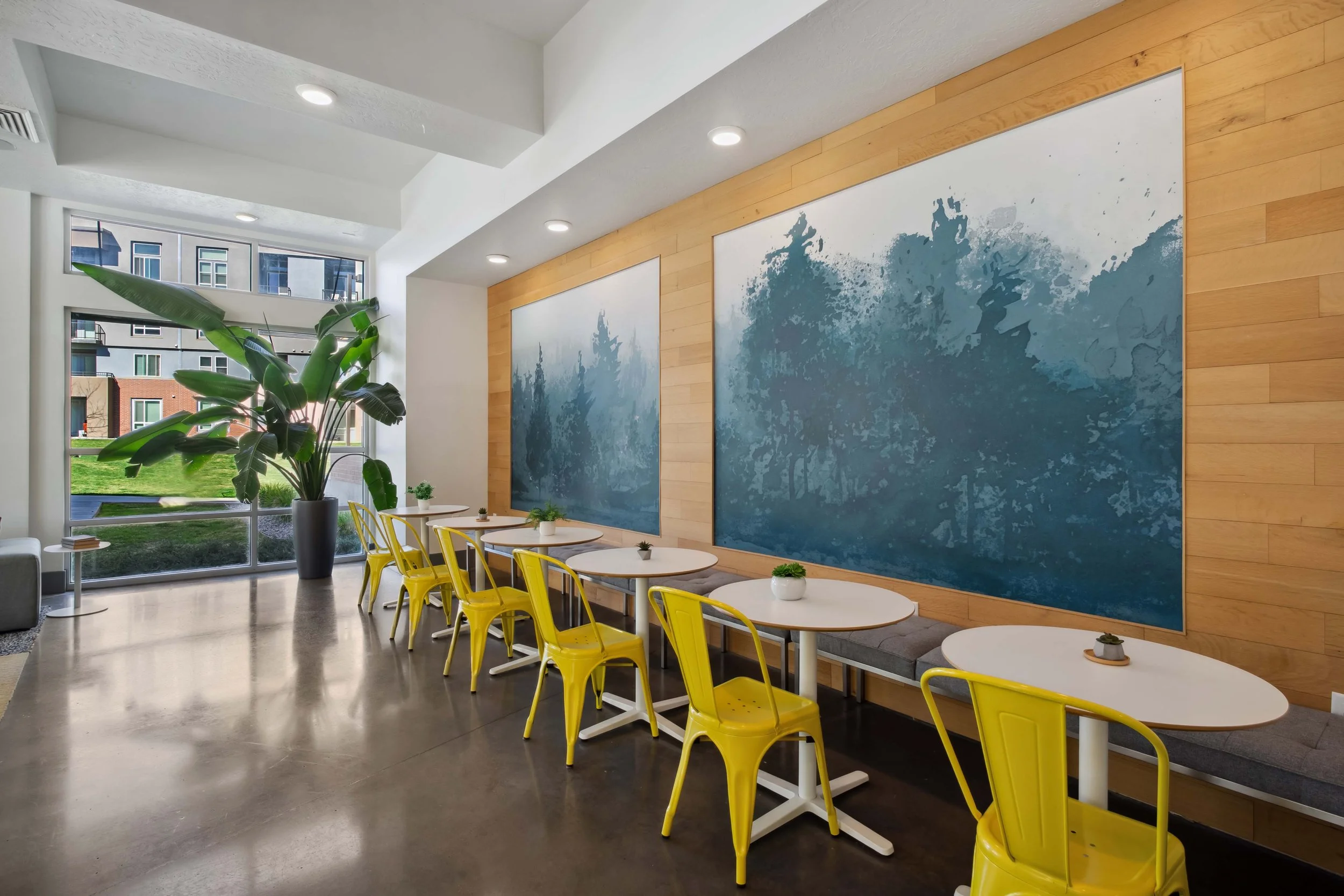 Modern café interior with yellow chairs, white tables, large green plant, wood accent wall, and abstract blue artwork